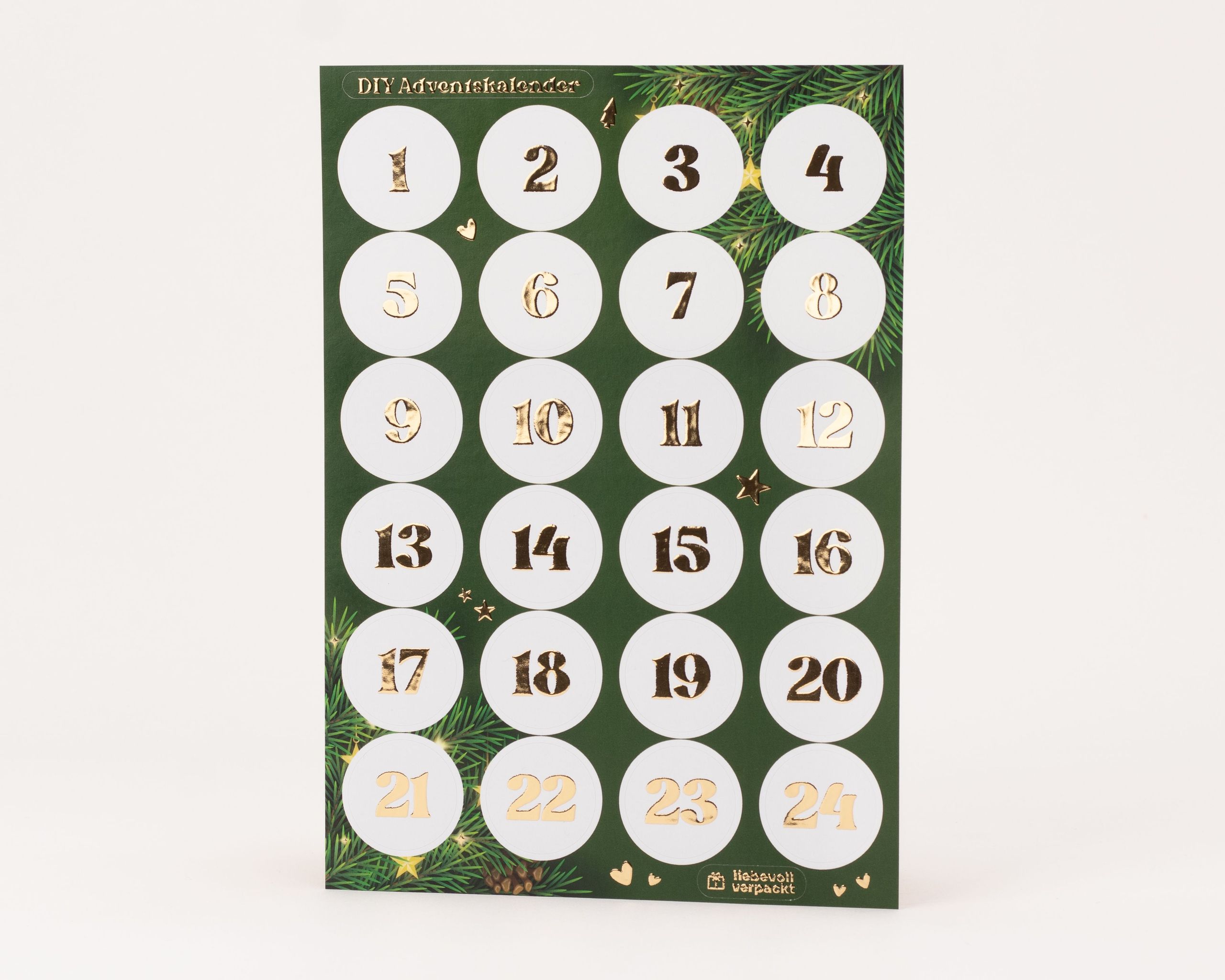 Eleganter Stickerbogen für einen DIY-Adventskalender mit 24 runden, weißen Aufklebern. Die Zahlen 1 bis 24 sind mit glänzender Goldfolie veredelt und heben sich vom dunkelgrünen Trägerpapier mit Tannenzweig-Motiven ab.