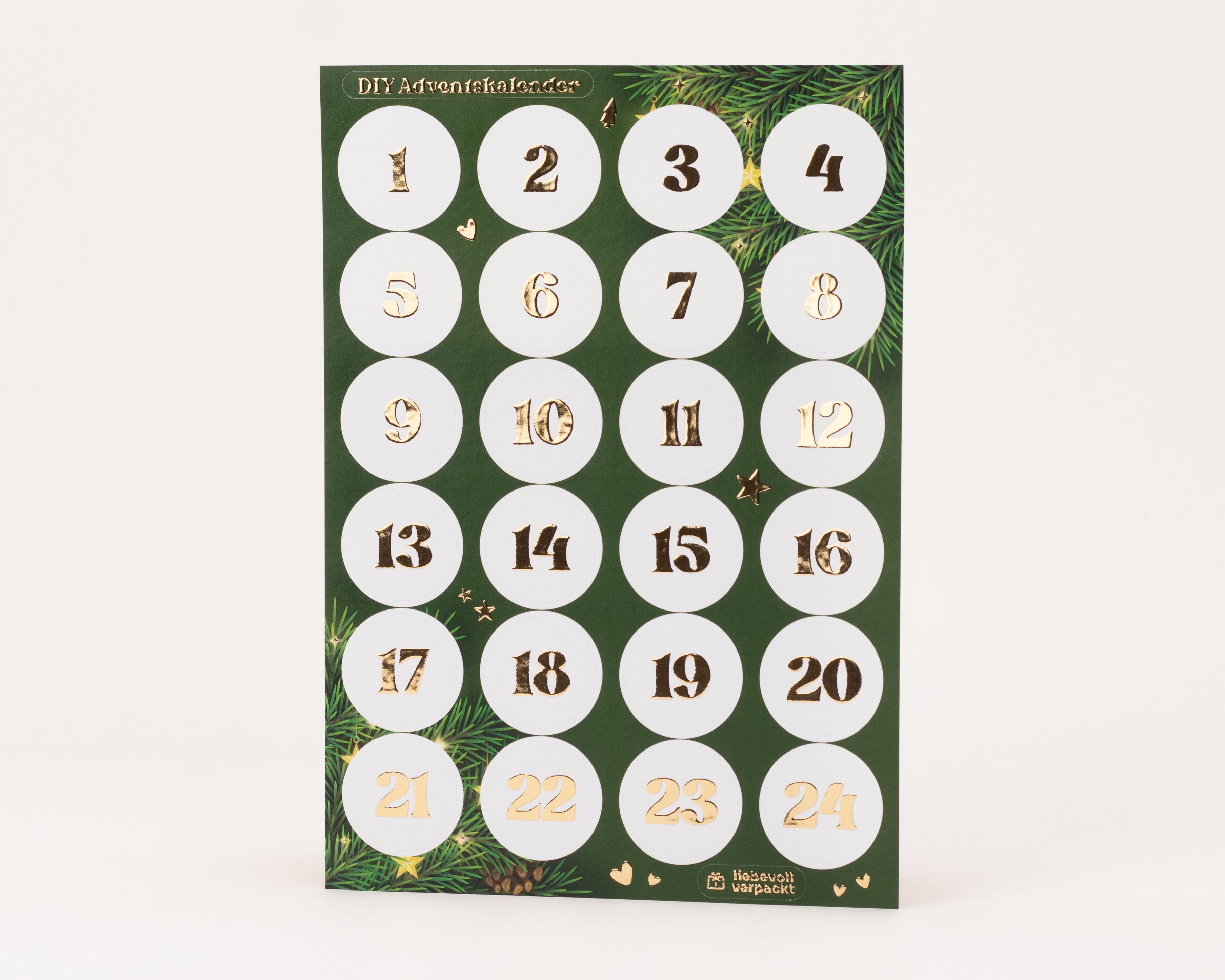 Eleganter Stickerbogen für einen DIY-Adventskalender mit 24 runden, weißen Aufklebern. Die Zahlen 1 bis 24 sind mit glänzender Goldfolie veredelt und heben sich vom dunkelgrünen Trägerpapier mit Tannenzweig-Motiven ab.