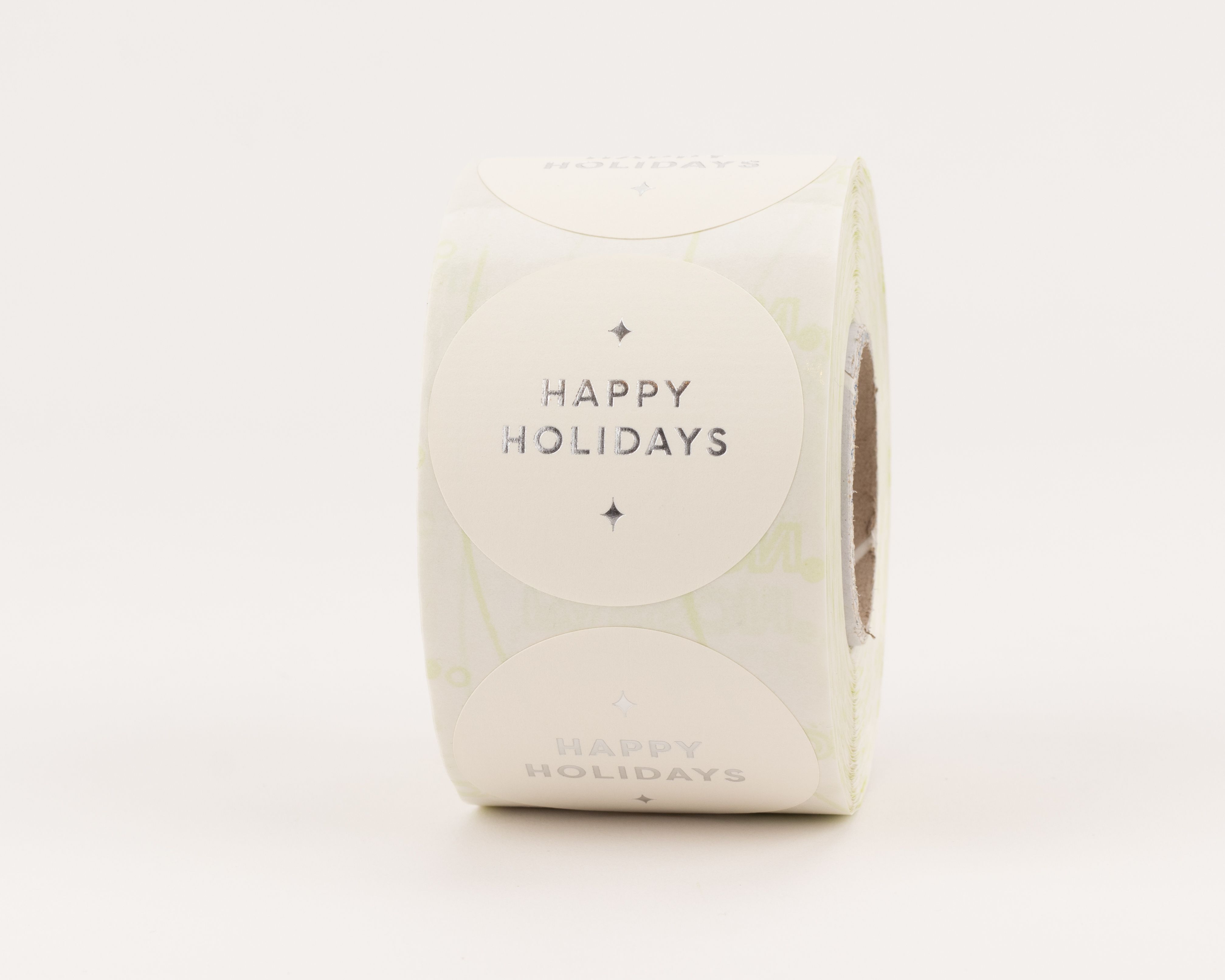 Aufkleber zeigen das Design Happy Holidays mit silberner Prägung auf cremeweißem Grund und kleinen sternförmigen Details