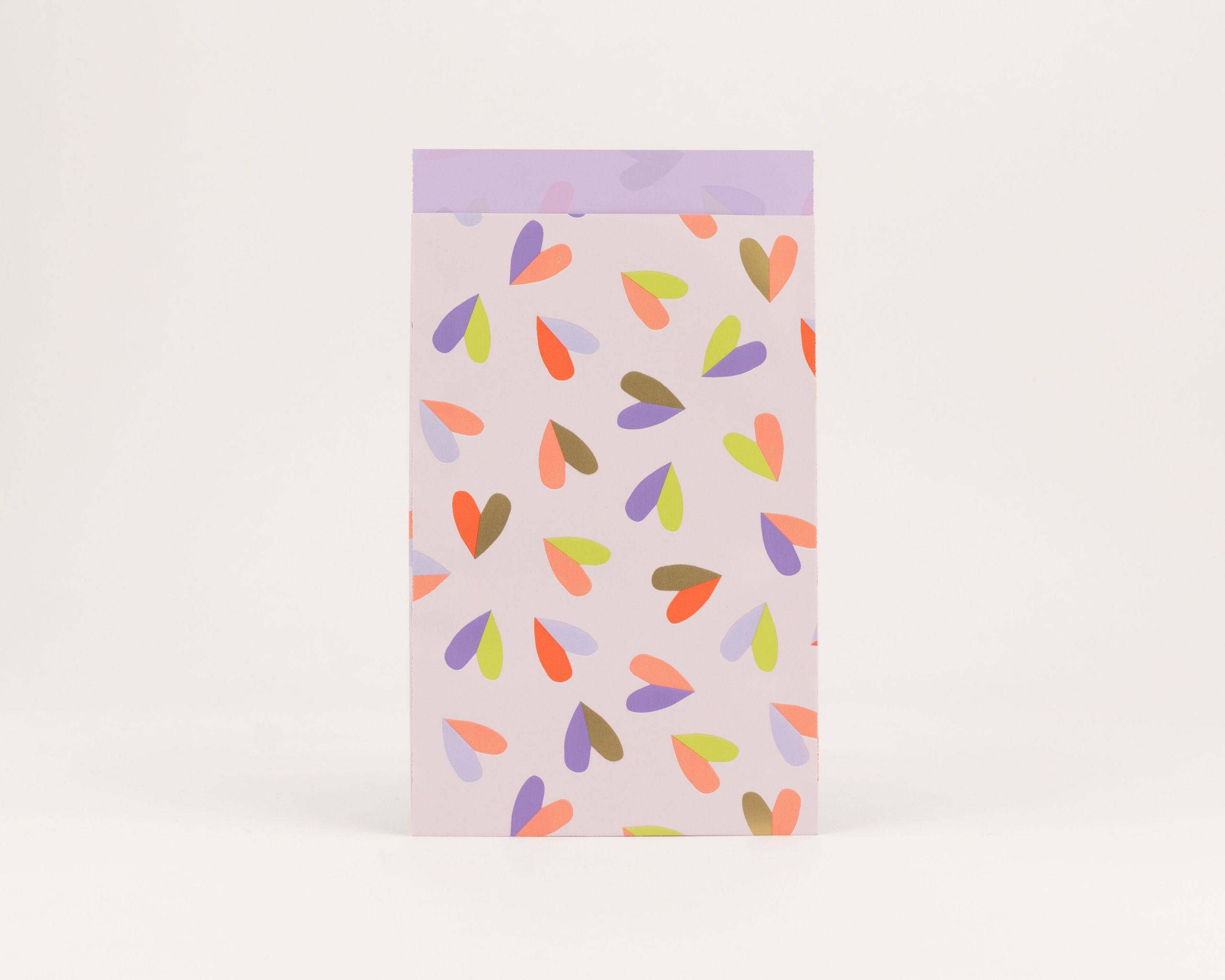 Flachbeutel zeigen ein Herzflatter-Muster auf hellviolettem Grund, mit Herzen in Orange, Violett und Limettengrün. Die Innenseite ist Fliederfarben