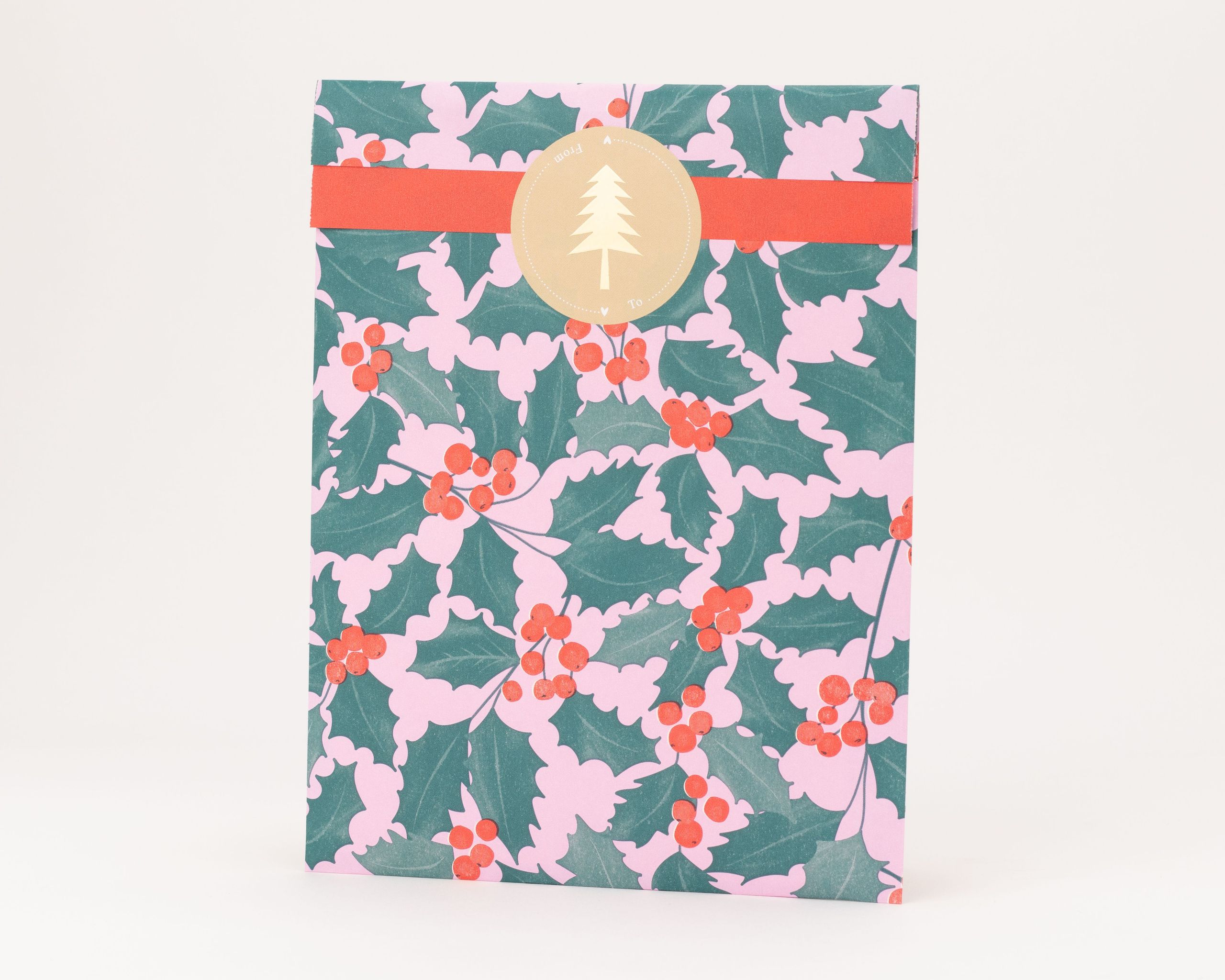 Flachbeutel mit einem rosa-grünen Stechpalmenmuster und roten Beeren auf der Außenseite, zusätzlich mit einem hellbraunen Tannenbaumaufkleber verziert
