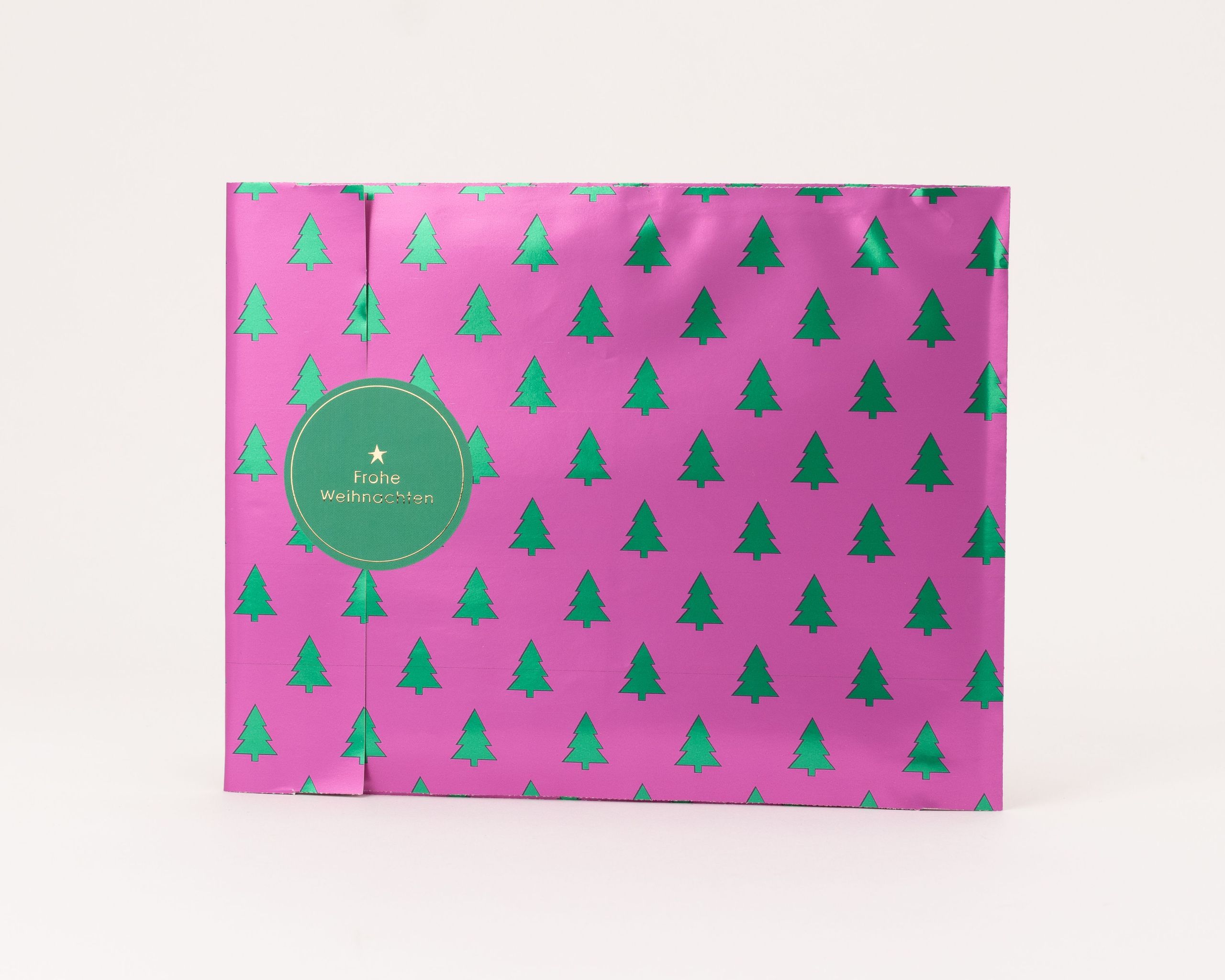 Flachbeutel in Pink mit metallic-grünem Tannenbaum-Muster. Ein runder grüner Weihnachtsaufkleber mit Goldprägung. Innenseite beige