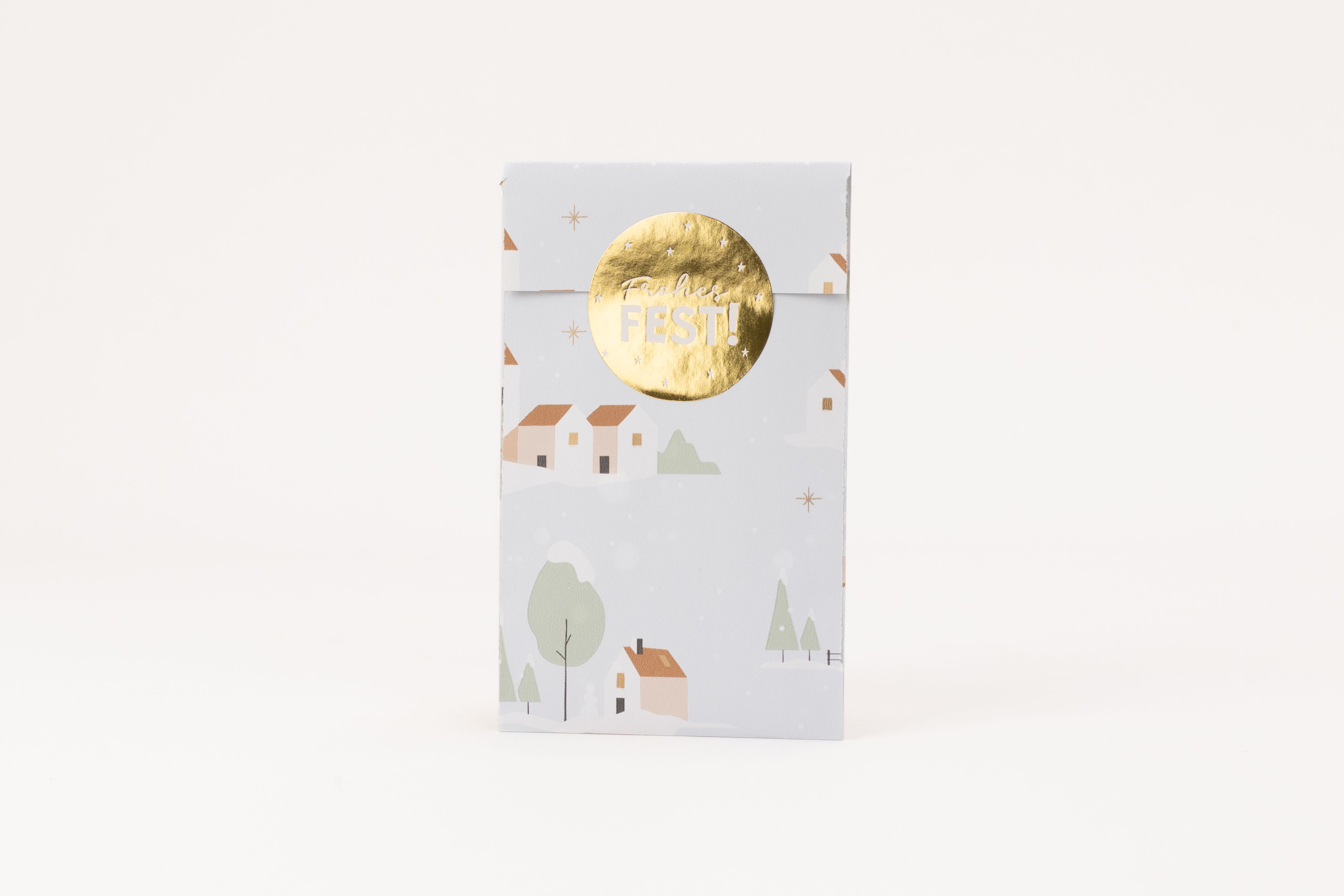 Flachbeutel mit hellblauem Winterdorfmotiv aus Häusern, Bäumen und Schnee. Goldene Sterne, Fenster und ein runder Goldsticker "Frohes Fest!"