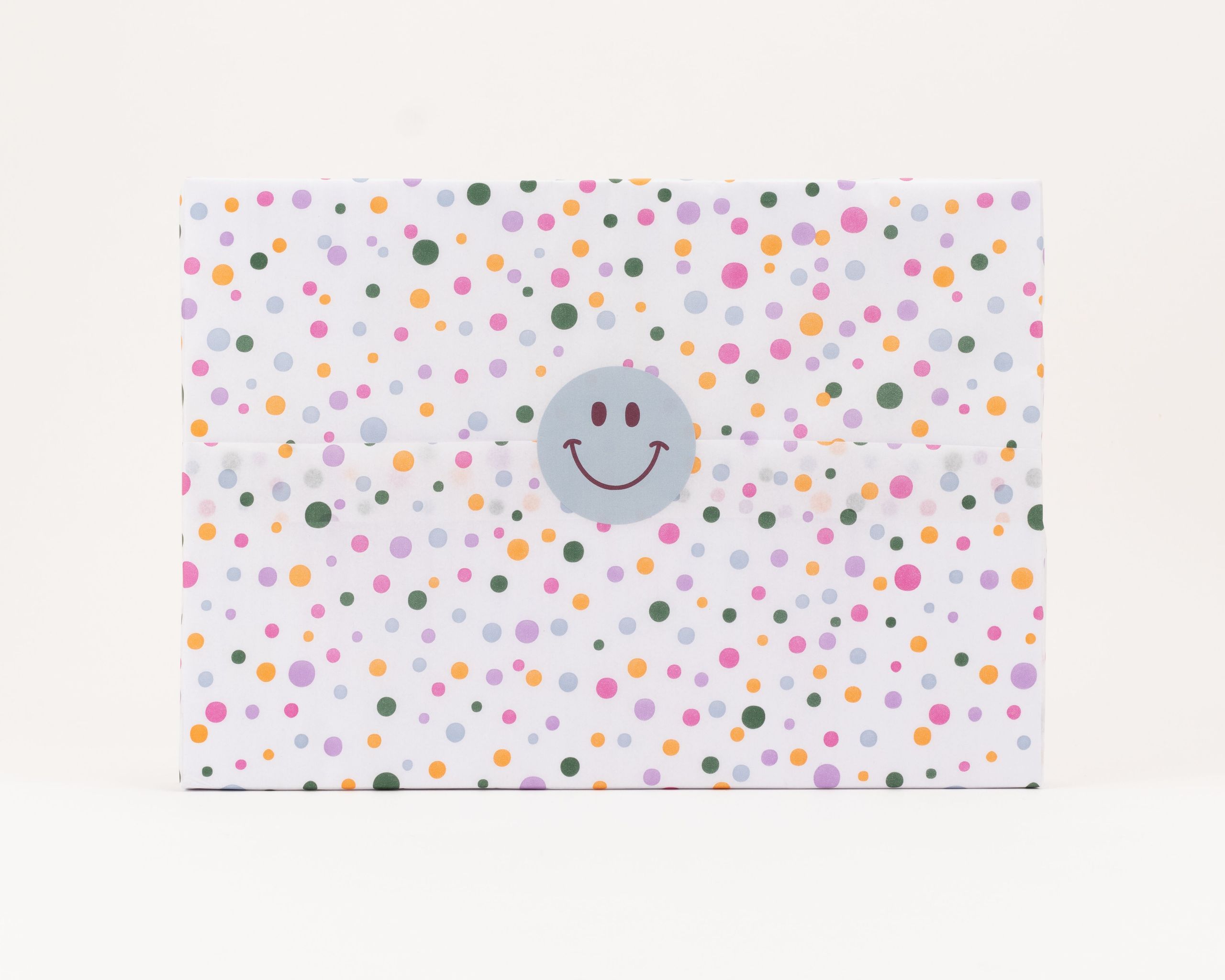 Seidenpapier in Weiß mit verschiedenfarbigen Konfettipunkten. Darauf ein runder hellgrauer Aufkleber mit einem braunen Smiley-Gesicht