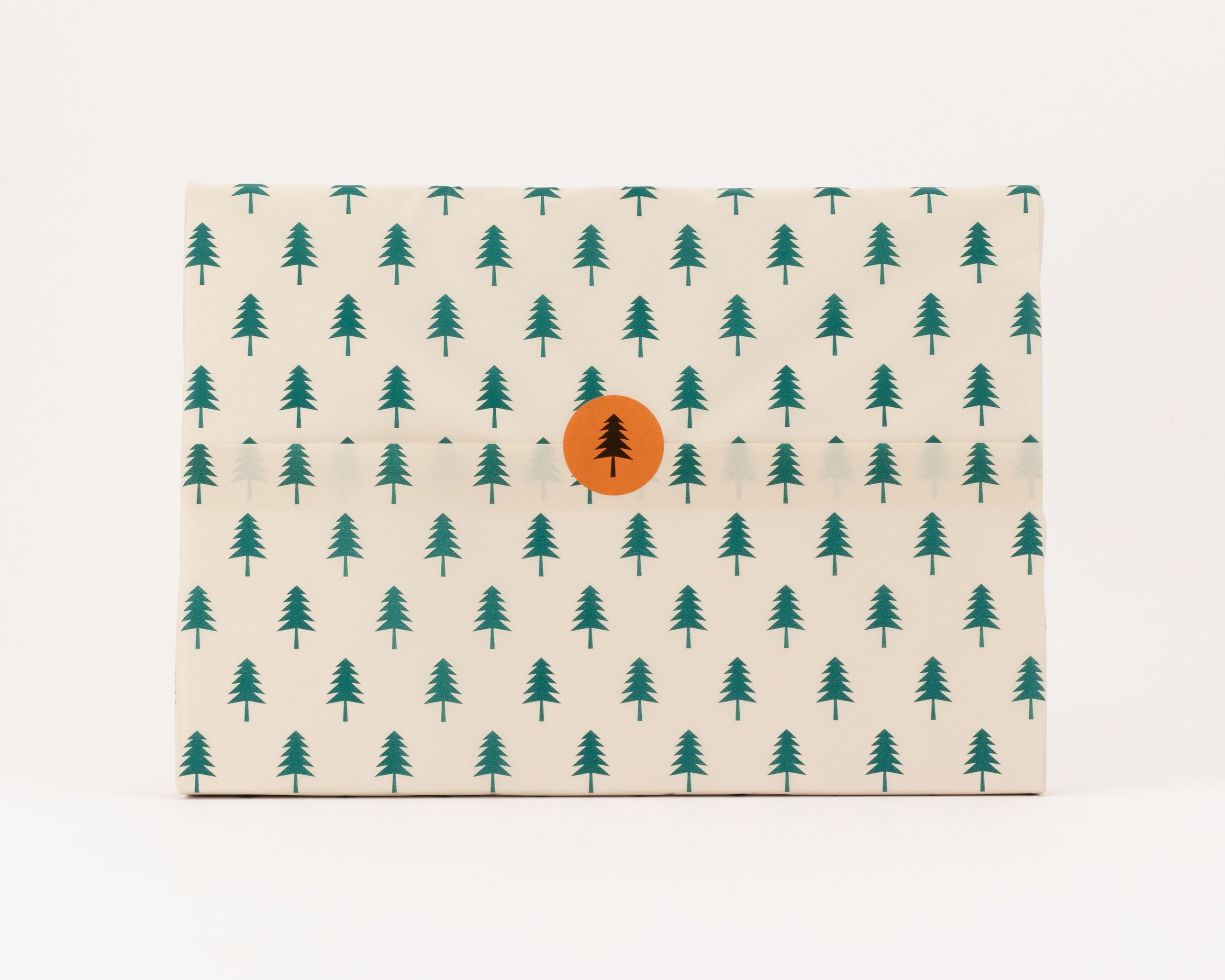 Seidenpapier mit beigem Hintergrund und einem Muster aus dunkelgrünen Tannenbäumen. Darauf ein runder, oranger Aufkleber mit einer Tannenbaum-Silhouette