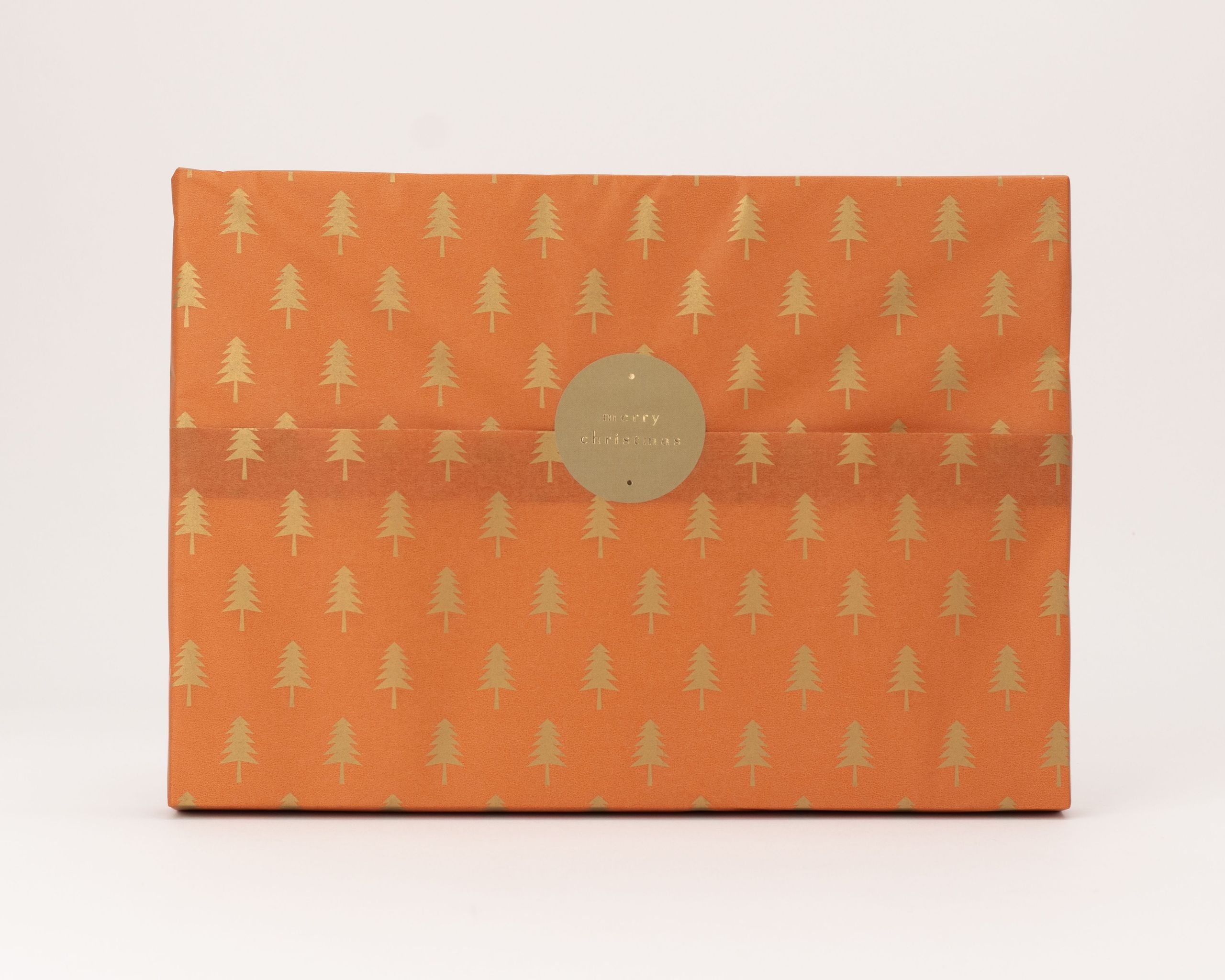 Seidenpapier in Rostfarbe mit goldenen Tannenbäumen als Muster. Darauf ein runder goldener Aufkleber mit dem Text merry christmas