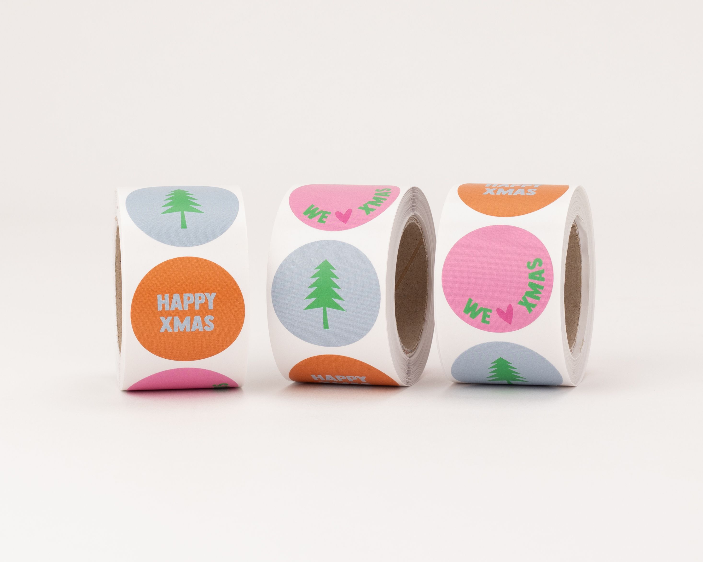 Aufkleber mit drei weihnachtlichen Motiven in Pink, Grün, Blau und Zimt. Runde Sticker mit Baum, 'HAPPY XMAS' und 'WE ❤️ XMAS' Designs