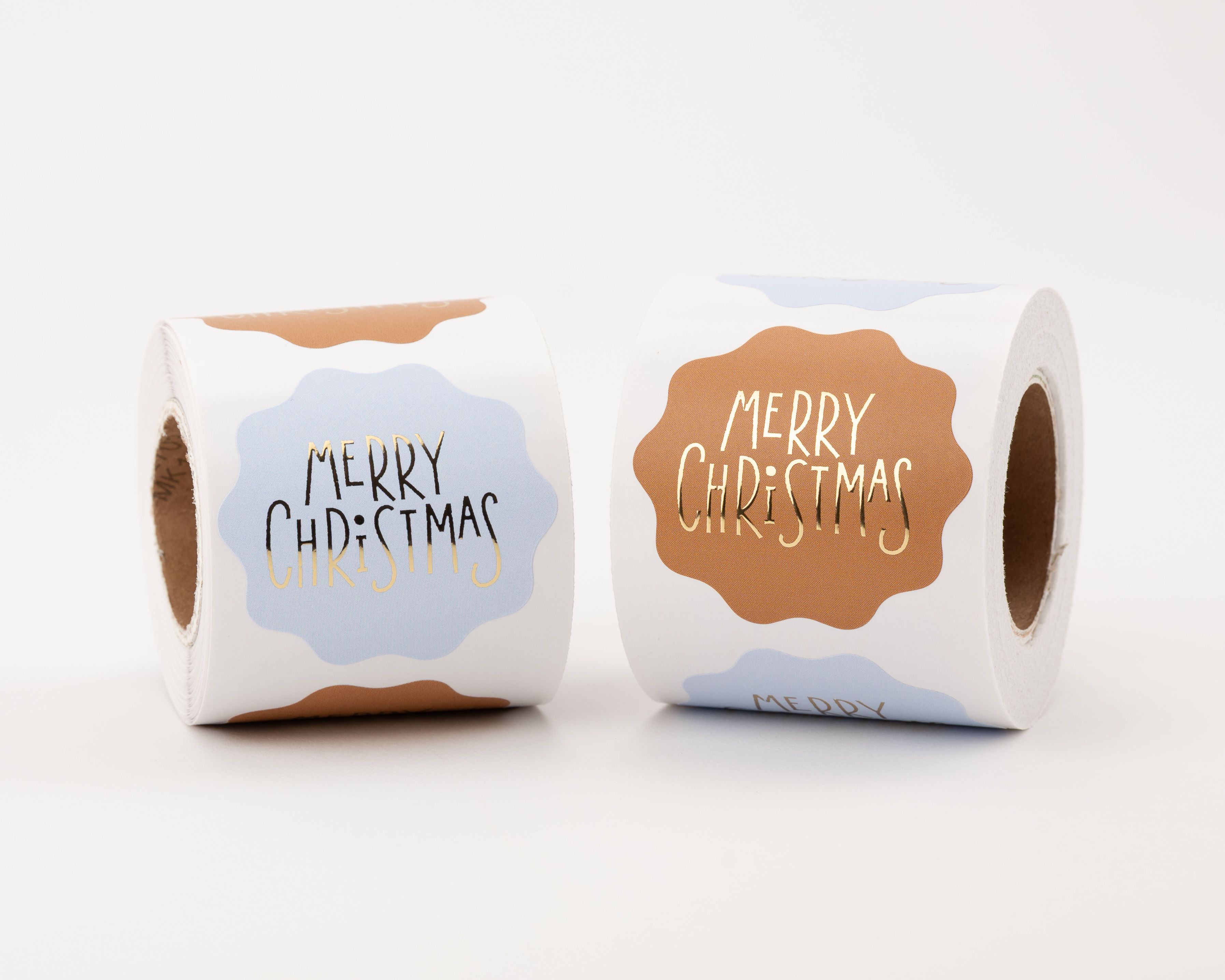Wellenförmige Aufkleber mit zwei Designs. Hellblaue Sticker zeigen 'Merry Christmas' in Schwarz und Gold, rostbraune Sticker mit goldener Schrift.