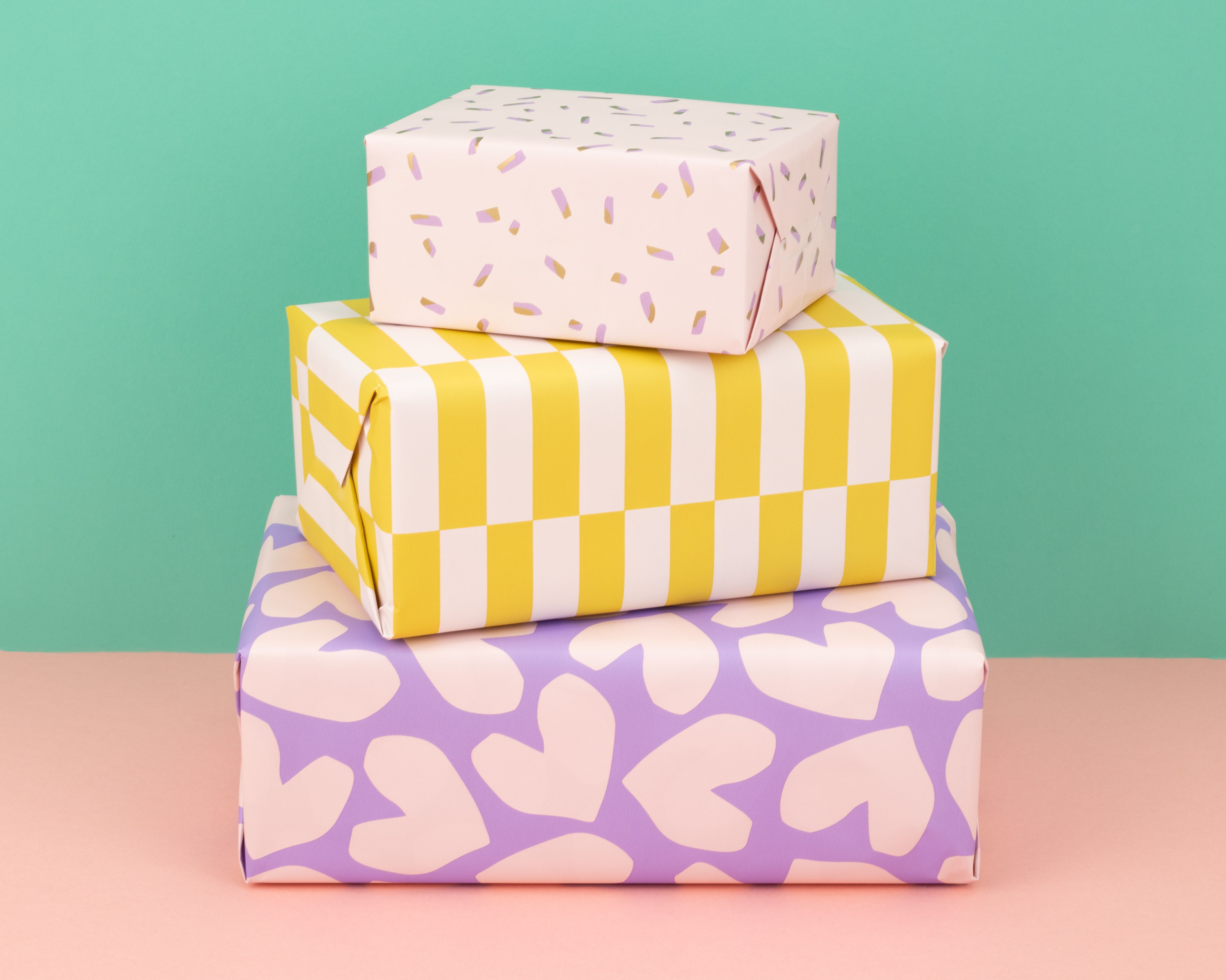 Geschenkpapier-Set mit drei gestapelten Paketen, verziert mit Papier in Flieder, Rosa und Gelb, mit Mustern aus Herzen, Blockstreifen und Konfetti