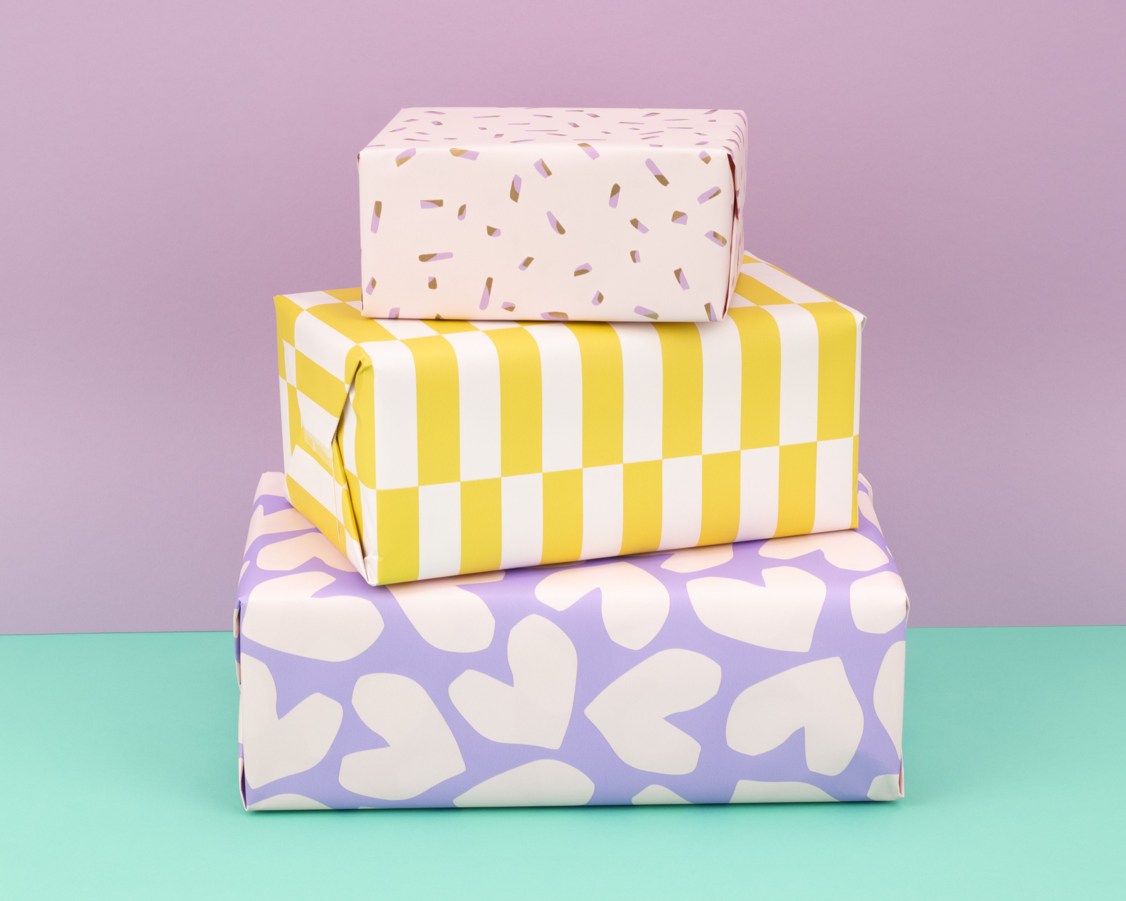 Geschenkpapier-Set mit drei gestapelten Geschenken in Flieder, Gelb und Creme mit Mustern wie Herzen, Blockstreifen und Konfetti