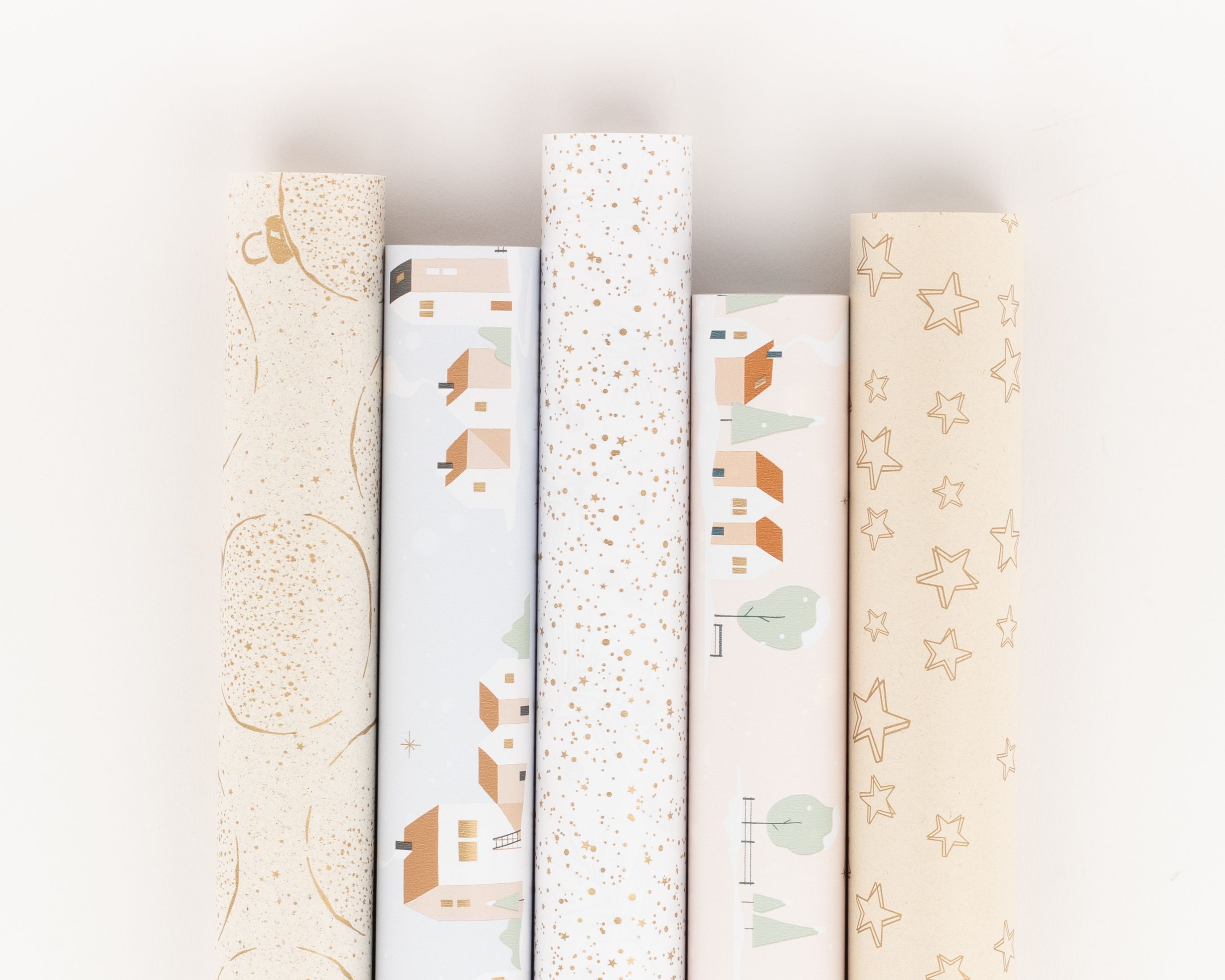 Geschenkpapier-Set mit fünf aufrecht stehenden Rollen in Pastellfarben, Beige und Weiß. Die Muster zeigen Winterdörfer, goldene Sterne und abstrakte Linien.