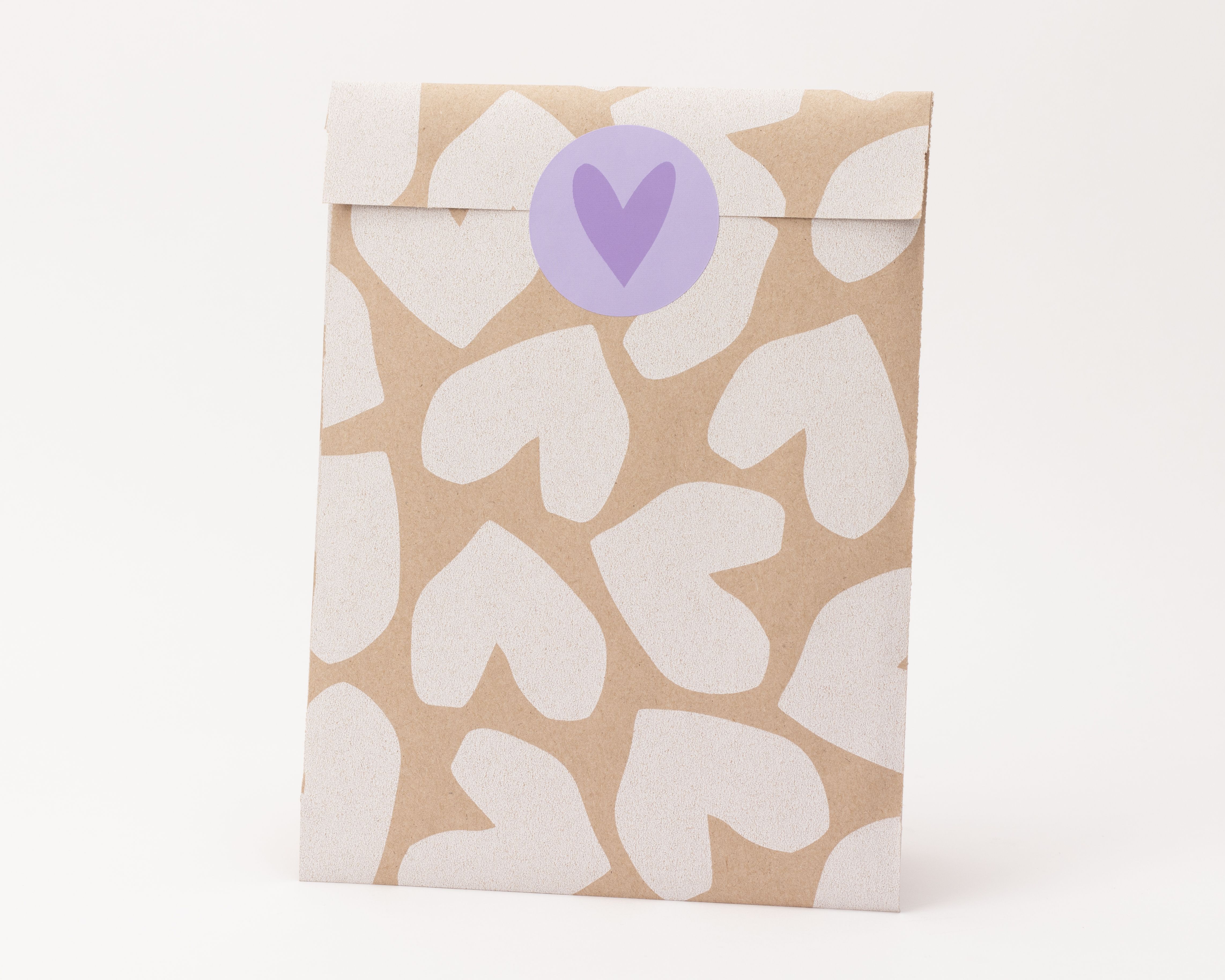 Flachbeutel aus Kraftpapier mit weißen Herzen als Muster und einem runden lila Aufkleber zum Verschluss