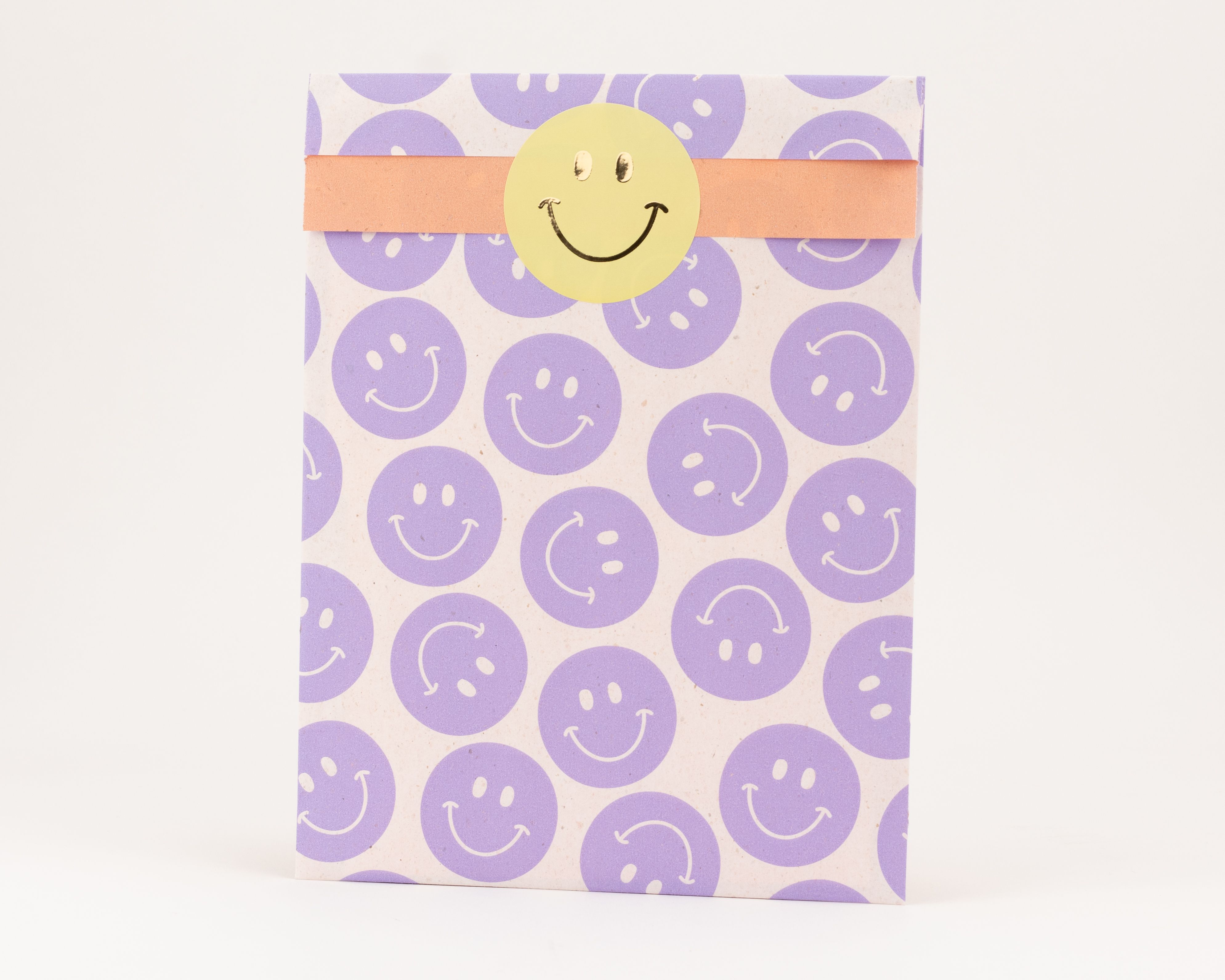 Flachbeutel mit fliederfarbenen Smileys und einer orangefarbenen Innenseite. Ein gelber Sticker mit schwarzem Smiley ist darauf zu sehen