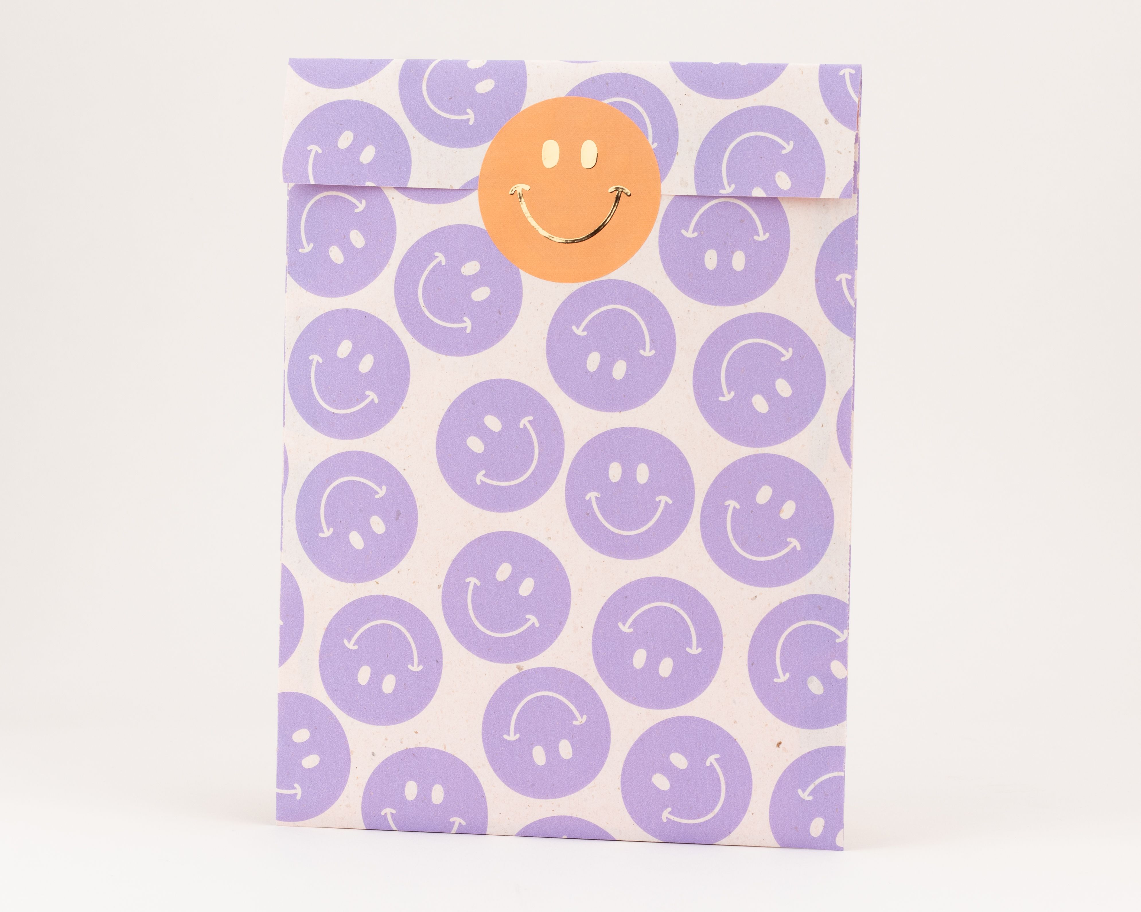 Flachbeutel mit einem Muster aus fliederfarbenen Smileys und einer hellen, unbedruckten Innenseite, verschlossen mit einem orangefarbenen Aufkleber mit goldenem Smiley