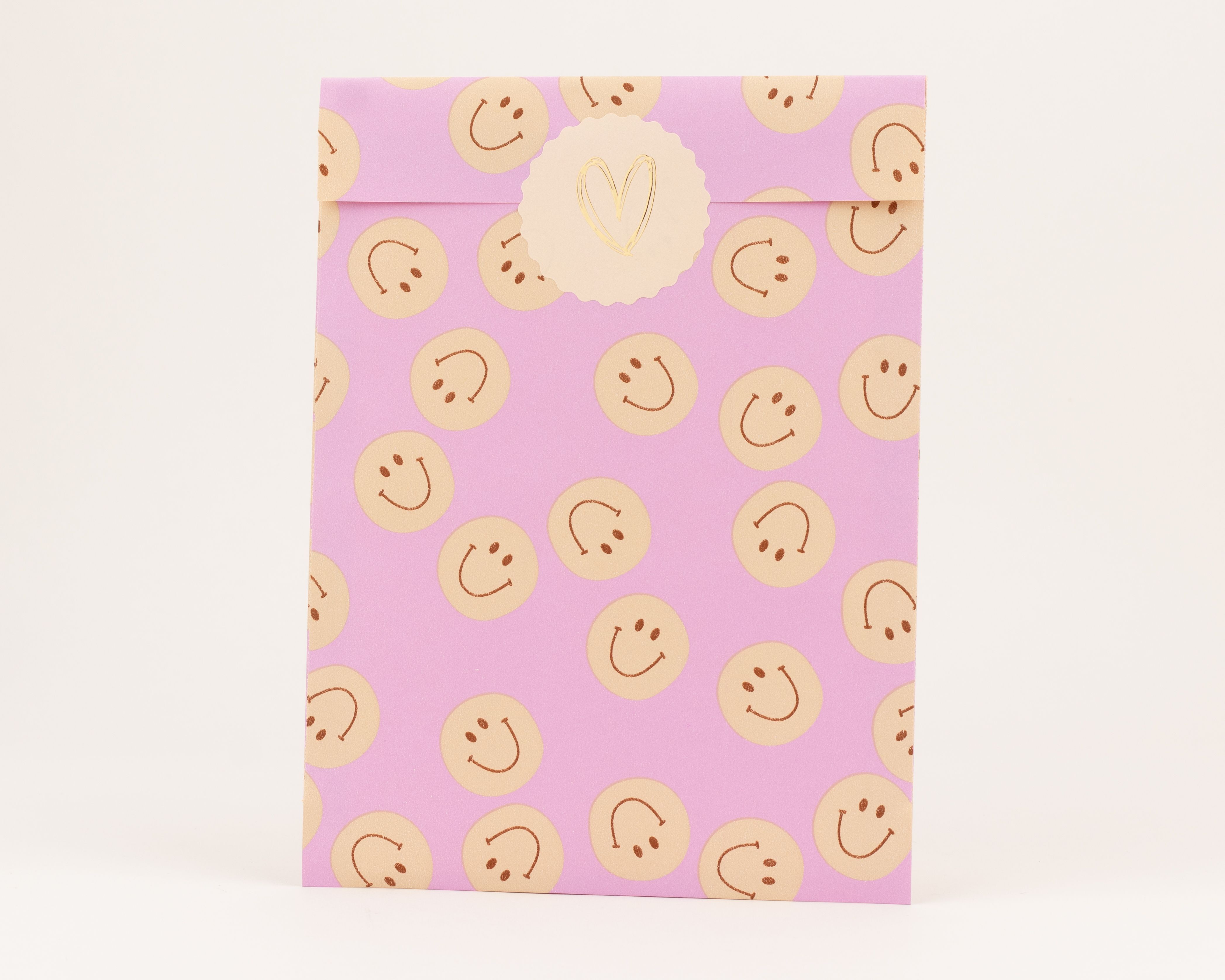 Flachbeutel in Flieder mit beigen Kreisen und orangefarbenen Smileys als Muster, verschlossen mit einem beigen Sticker mit goldenem Herz