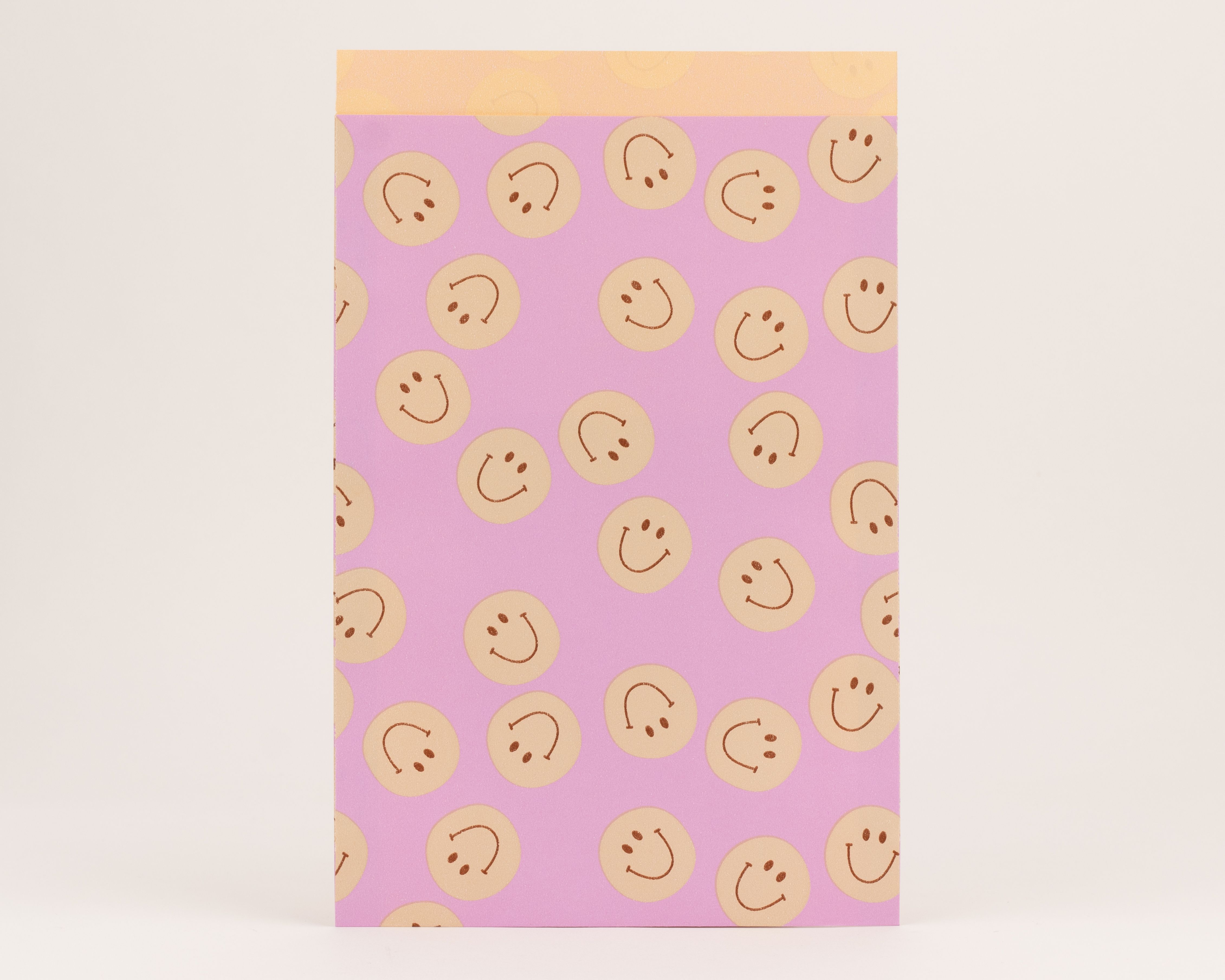 Flachbeutel mit fliederfarbener Außenseite und beigen Smileys. Die Innenseite ist orange mit dezenten orangefarbenen Smileys als Muster