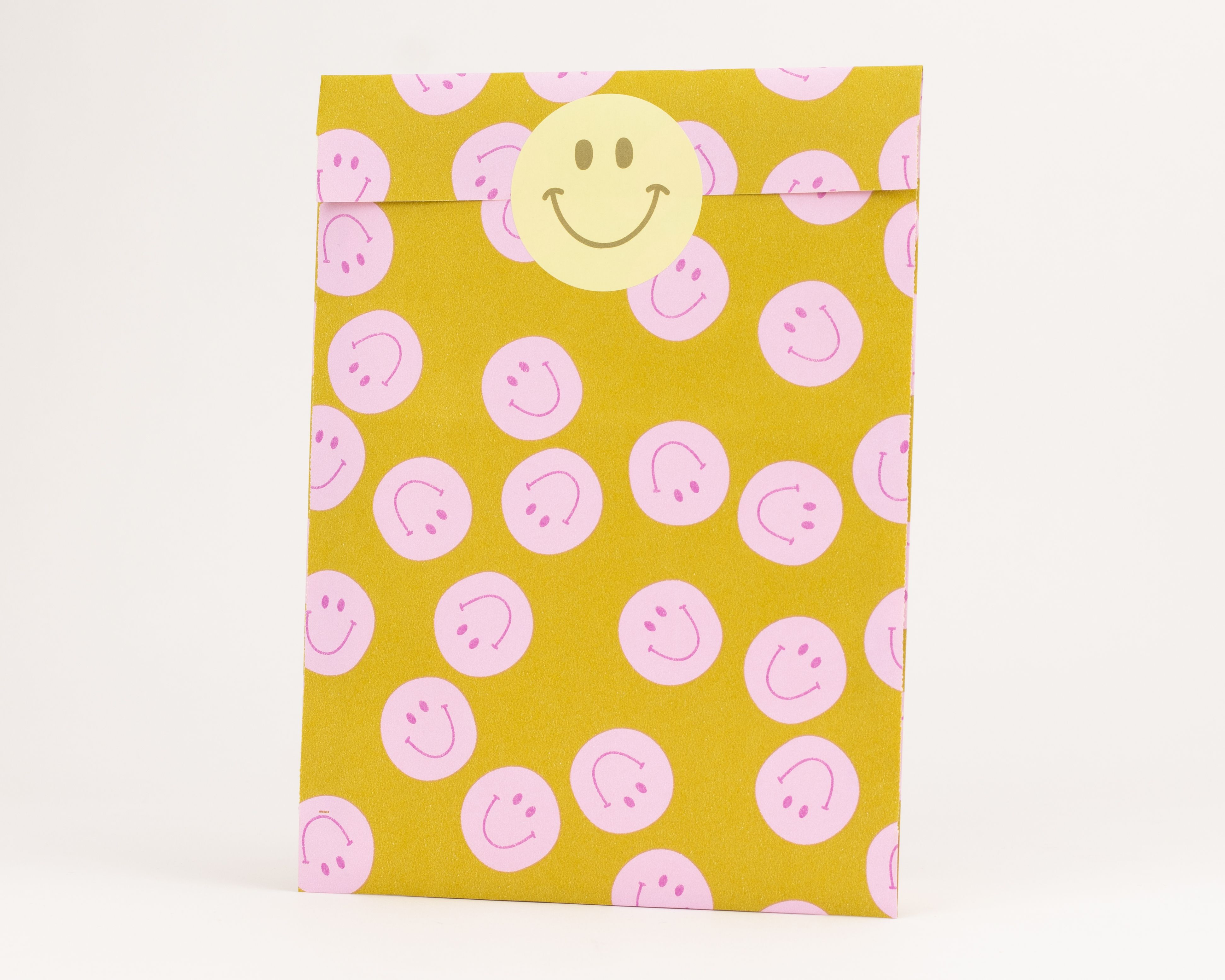 Flachbeutel in Gelb mit durchgehendem rosa Smiley-Muster und rosa Innenseite, mit einem runden gelben Smiley-Aufkleber