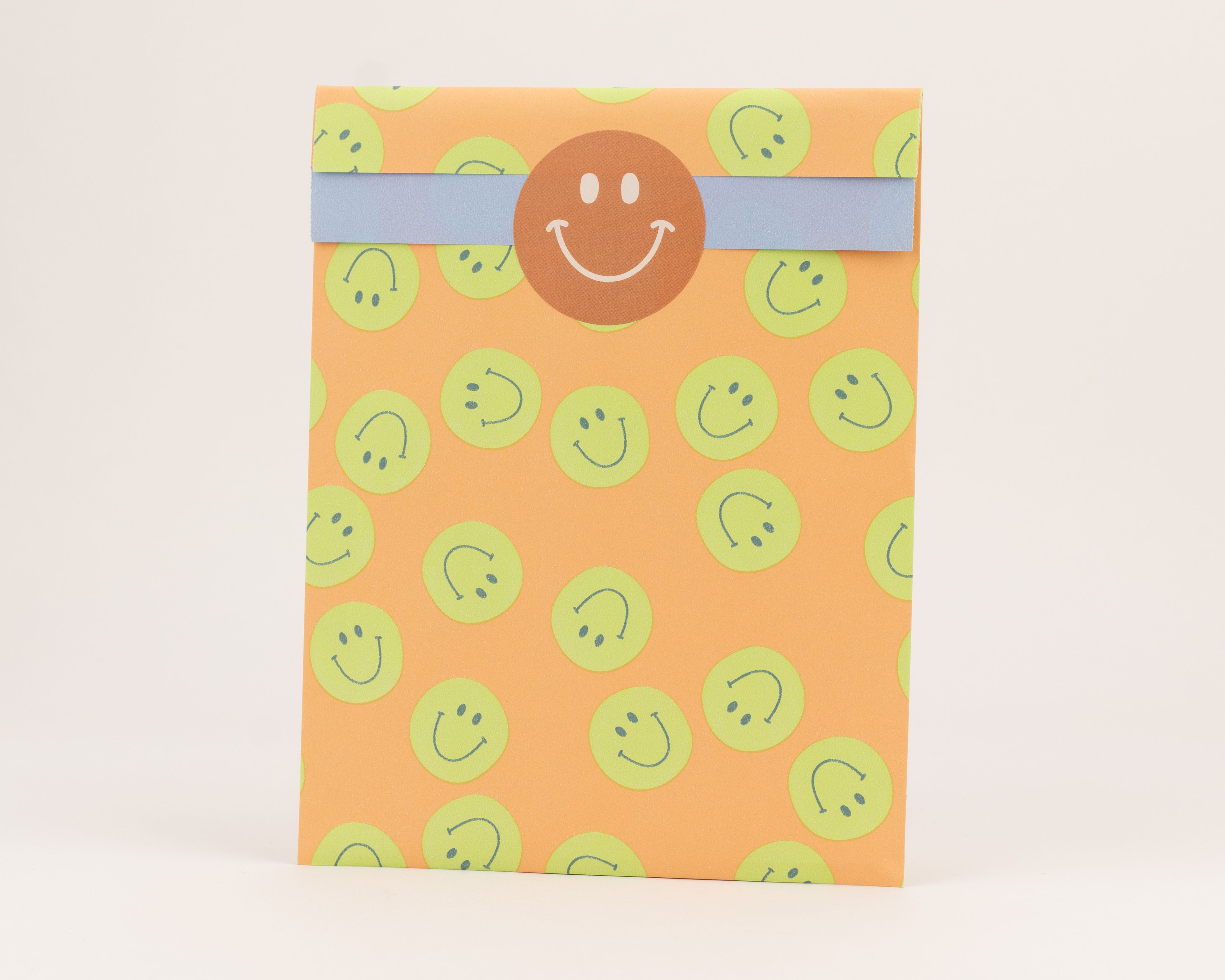 Flachbeutel mit orangefarbener Außenseite und grünen Smiley-Motiven. Die Innenseite ist blau, verdeckt von einem orangefarbenen Smiley-Aufkleber