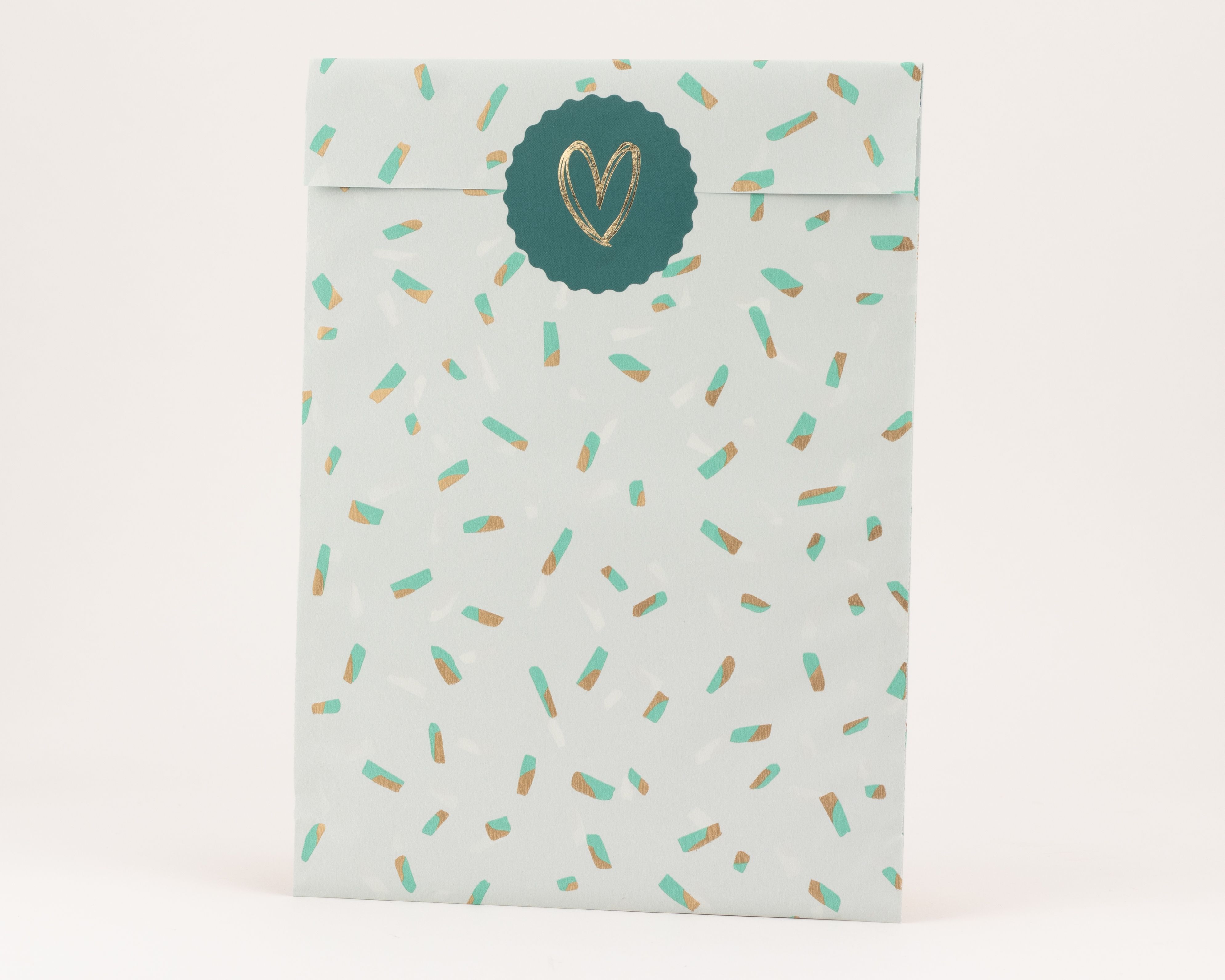 Flachbeutel in Salbeigrün mit einem Muster aus türkisen und goldfarbenen Sprinkles. Ein runder salbeigrüner Sticker mit einem Goldeffekt-Herz ist auf der Tüte platziert