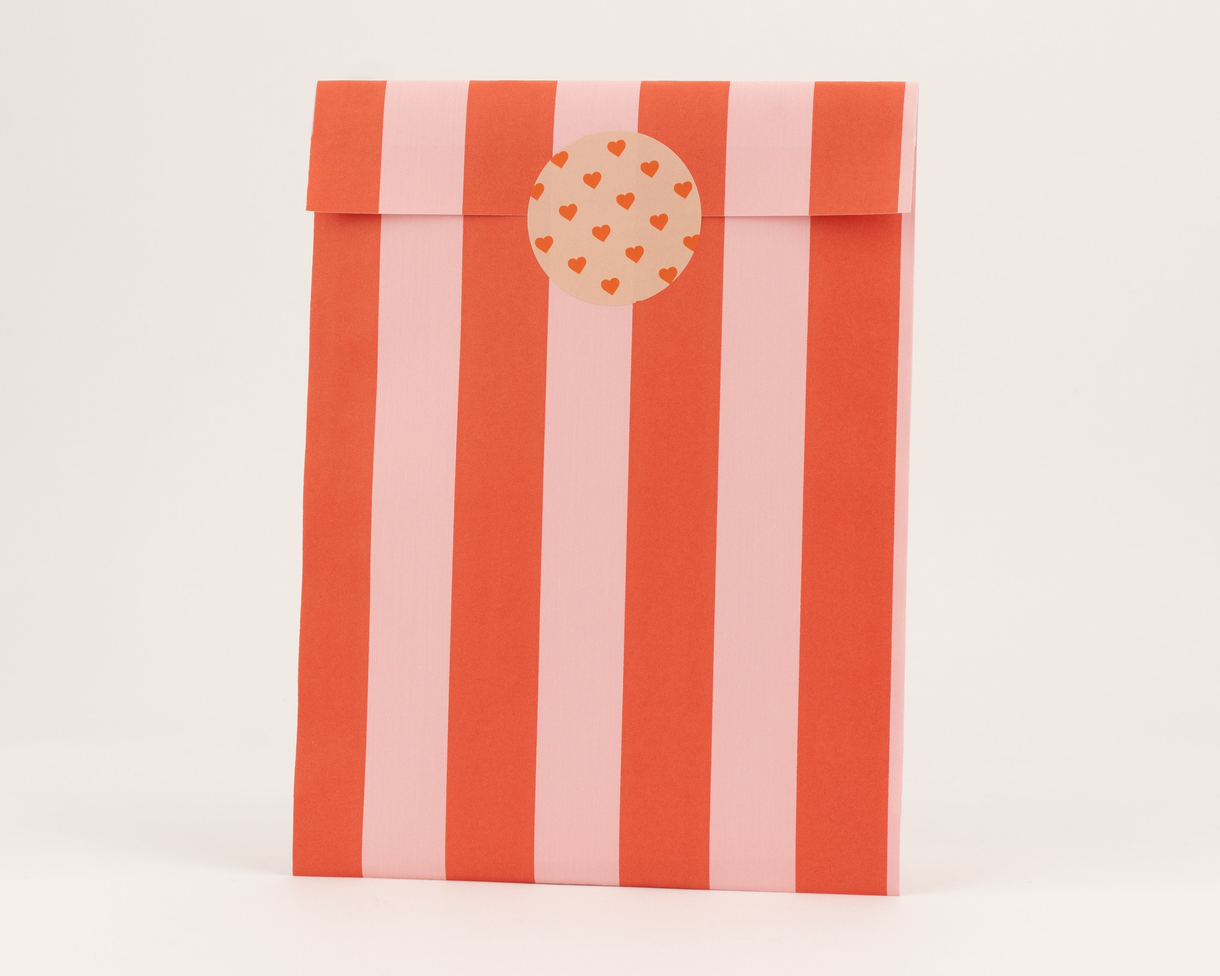 Flachbeutel mit roten und rosa Streifenmuster. Die Innenseite ist ebenfalls gestreift und mit einem Herz-Sticker verschlossen.