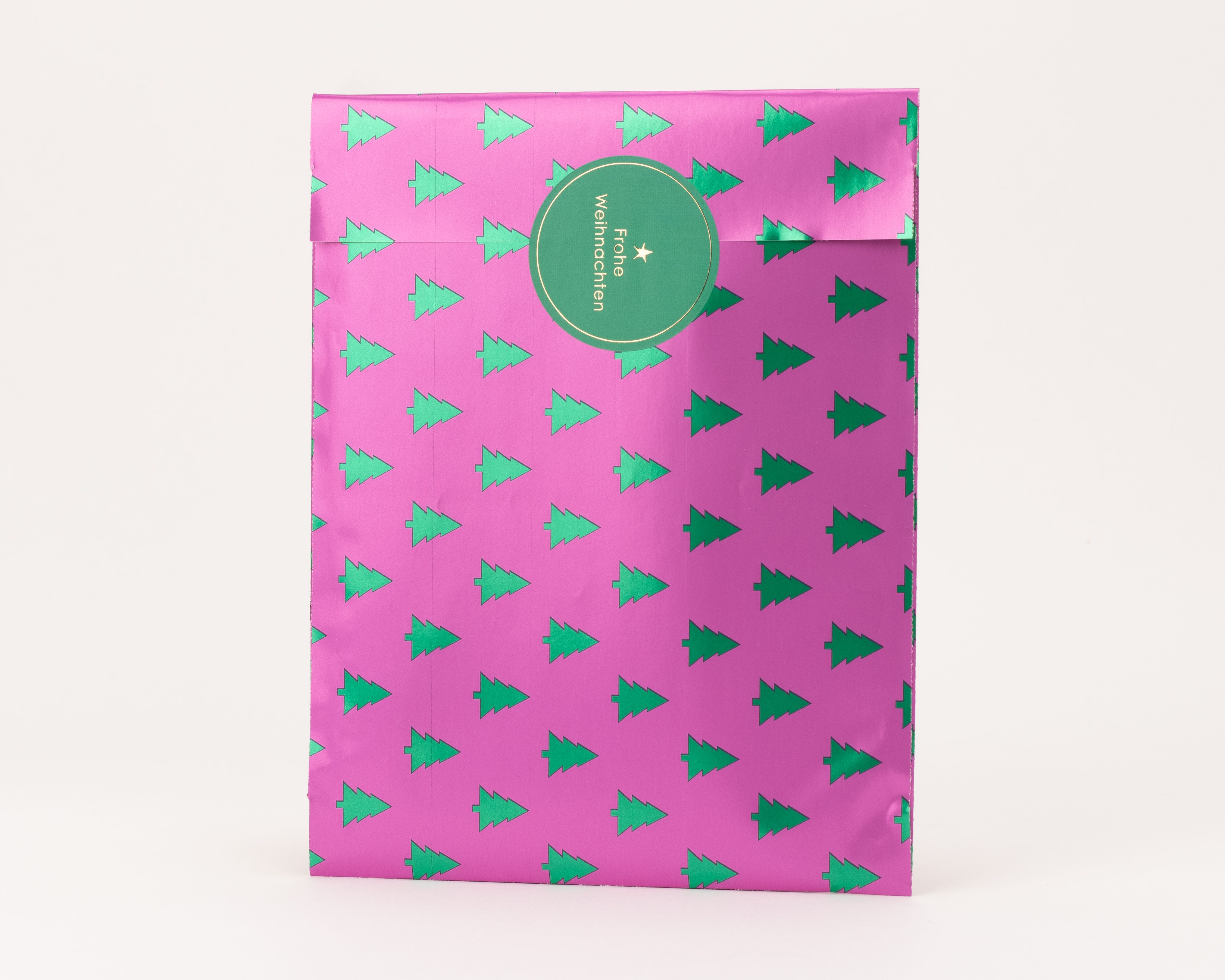 Flachbeutel in Pink mit metallisch grünen Tannenbäumen. Ein runder grüner Aufkleber mit 'Frohe Weihnachten' verschließt den Beutel