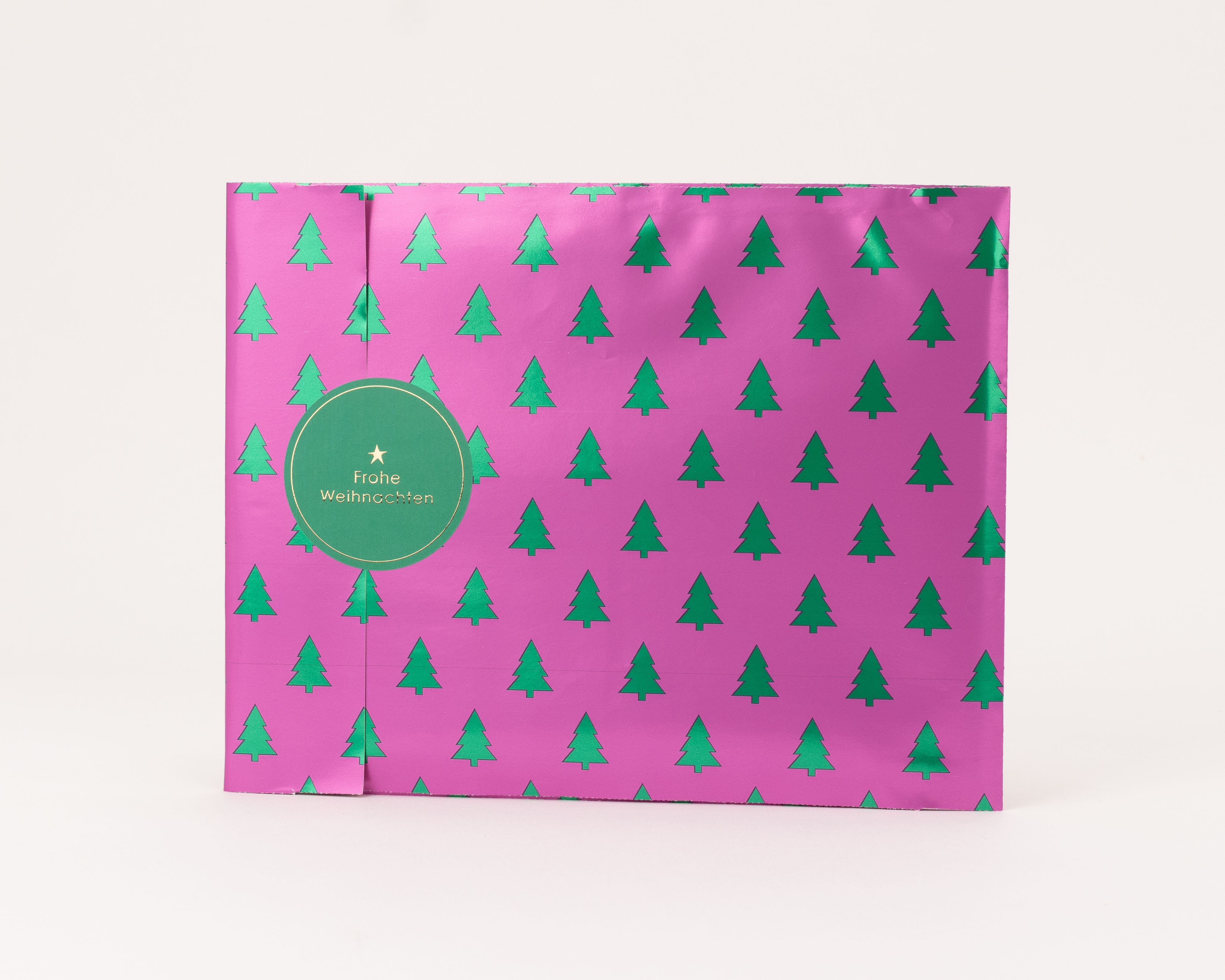 Flachbeutel in Pink mit metallic-grünem Tannenbaum-Muster. Ein runder grüner Weihnachtsaufkleber mit Goldprägung. Innenseite beige