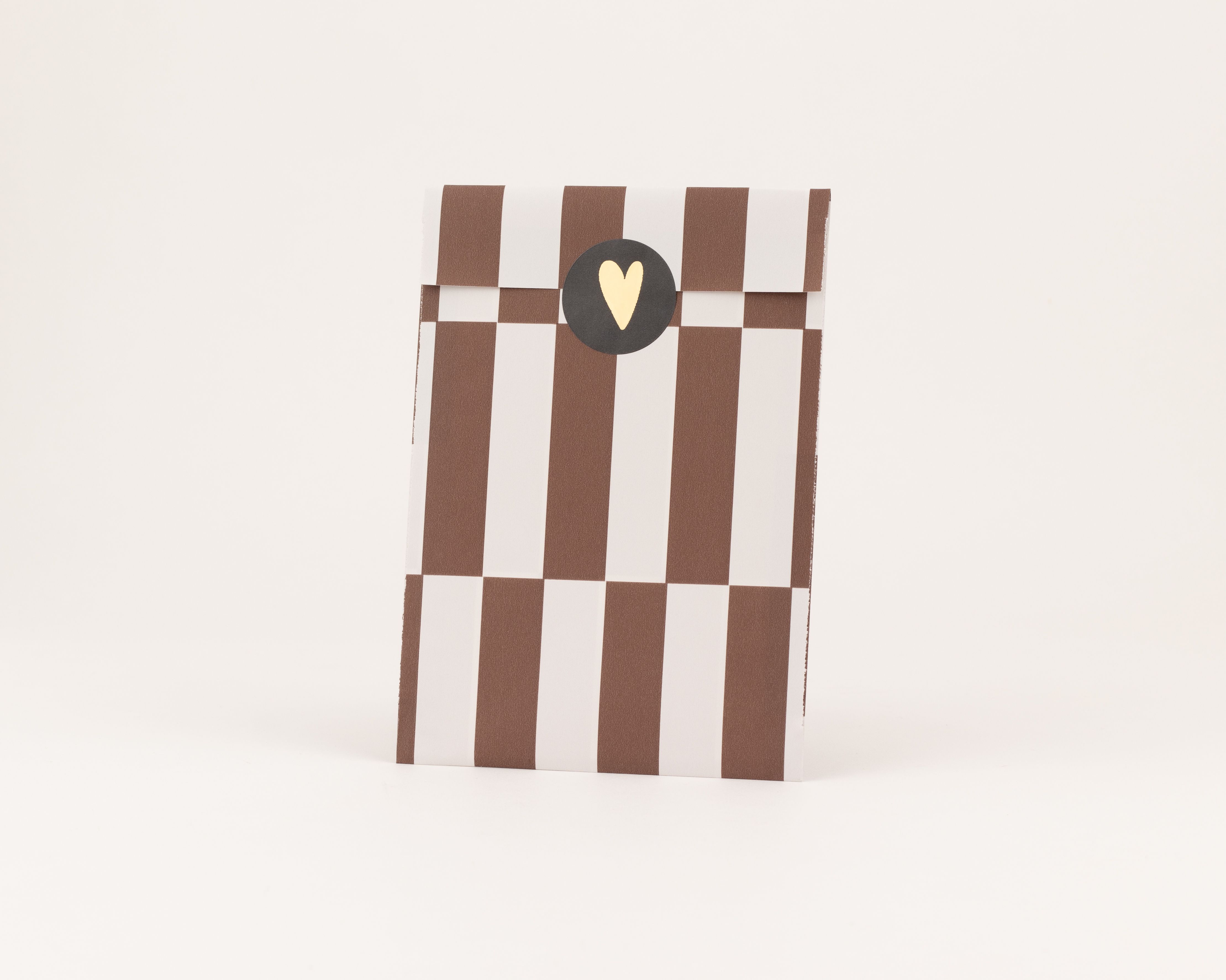 Flachbeutel mit braun-weißen Streifen und einer Innenseite mit braun-weißem Karomuster, verschlossen mit einem schwarzen Sticker und goldenem Herz