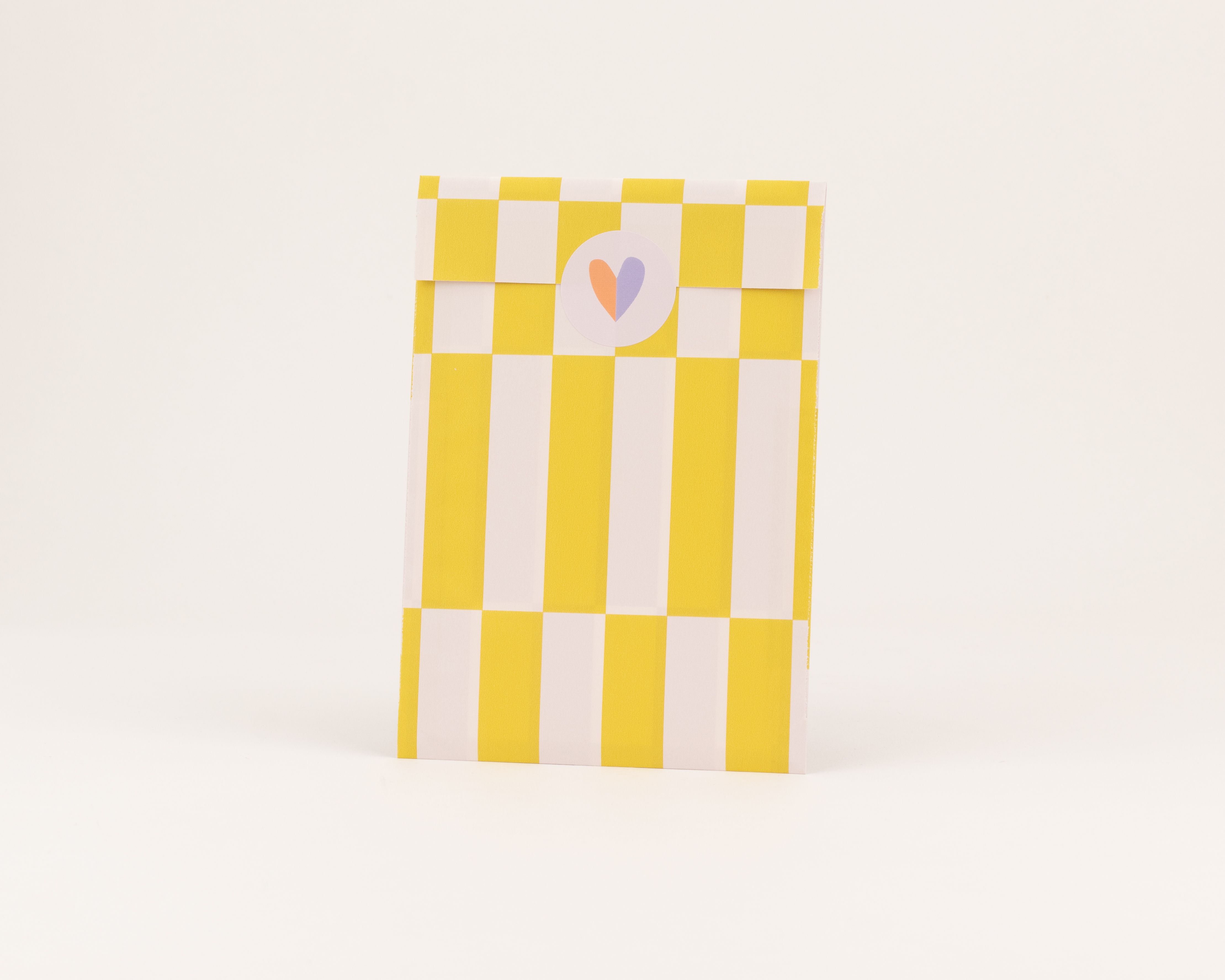 Flachbeutel mit auffälligem gelb-weiß gestreiftem Muster und karierter Innenseite, verziert mit einem Sticker mit lilafarbenem Herz