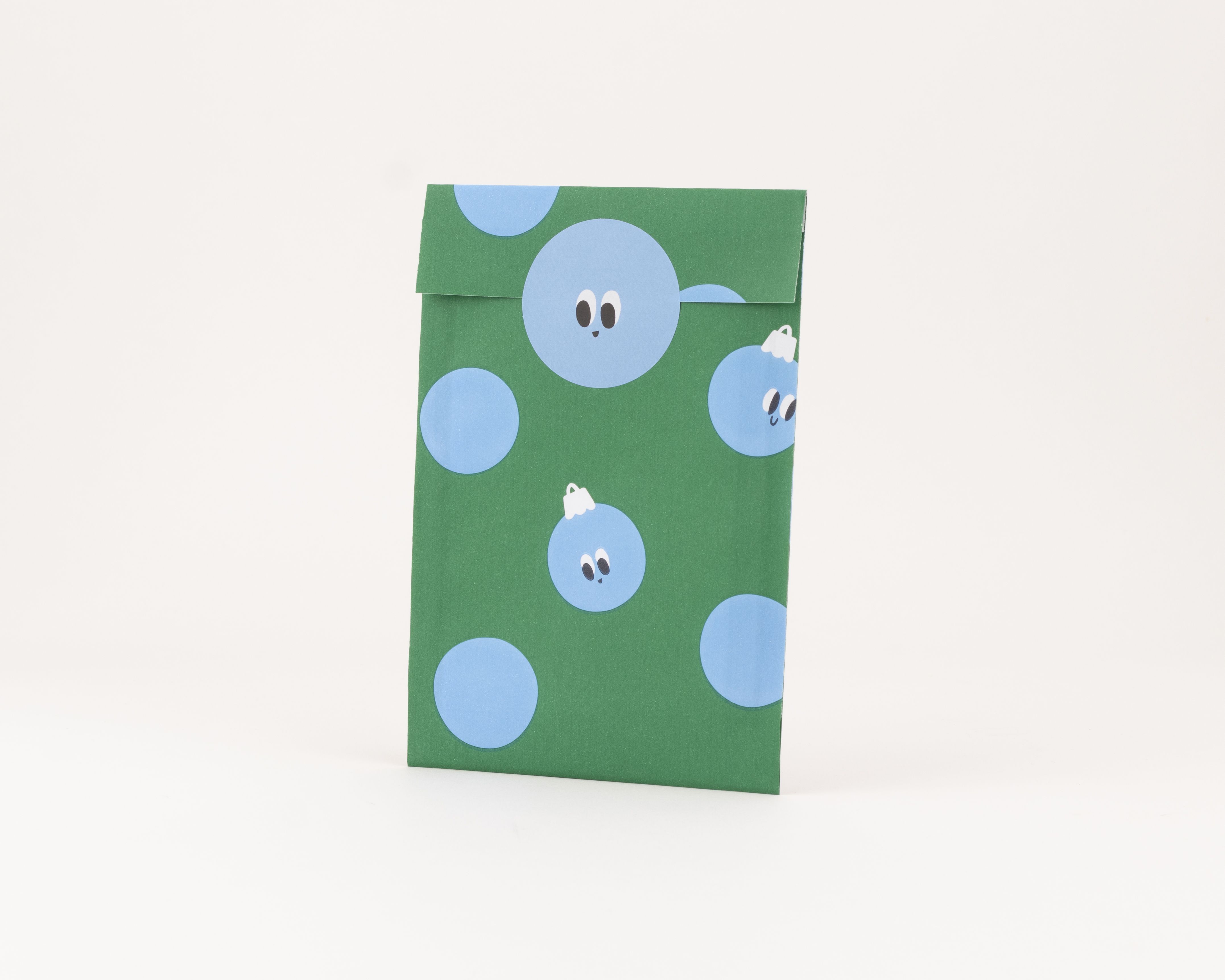 Flachbeutel in Grün mit einem verspielten Muster aus blauen Kugeln, von denen einige Gesichter als Weihnachtskugeln zeigen
