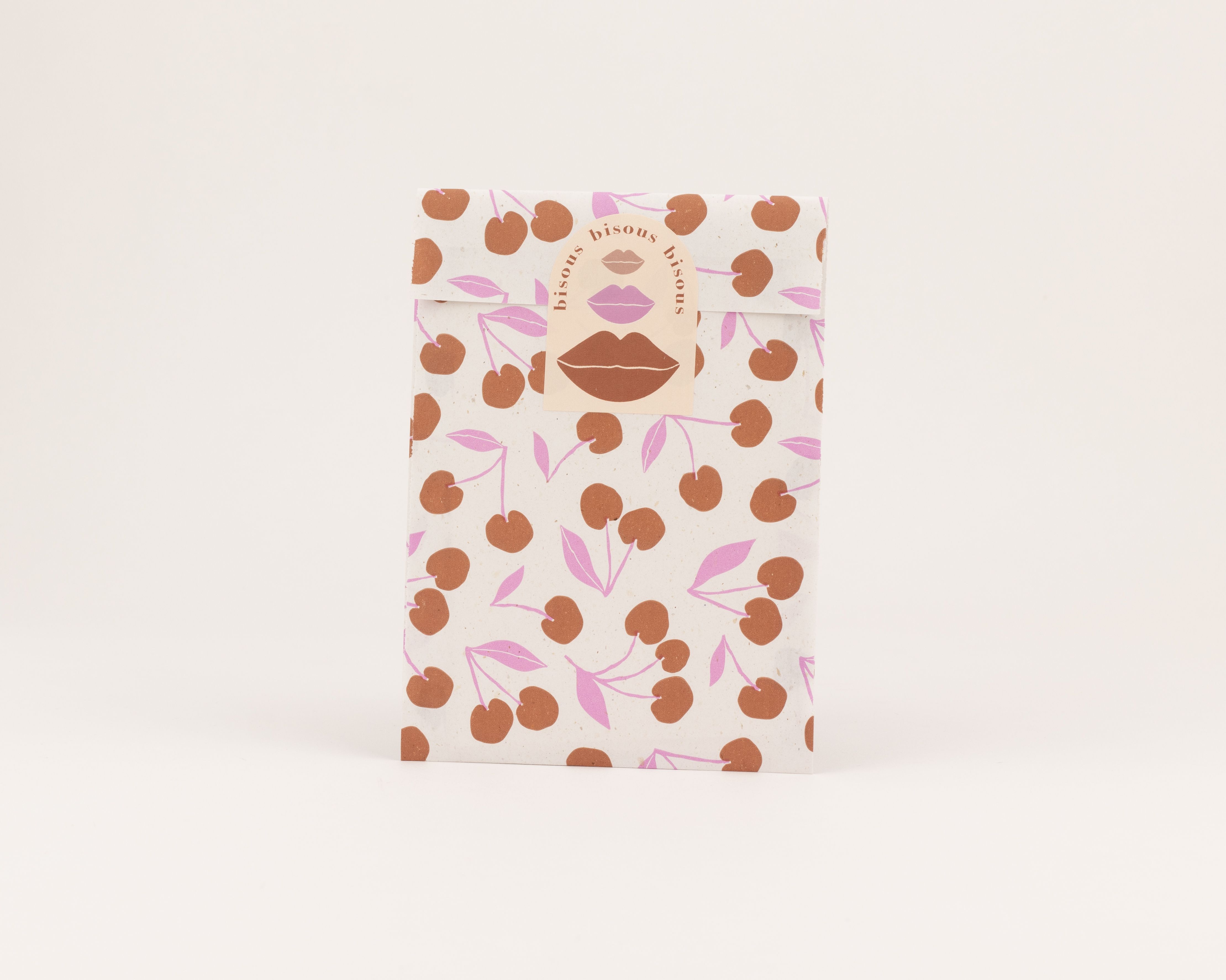 Flachbeutel mit Muster aus braunen Kirschen und pinken Blättern. Die Innenseite zeigt einen Aufkleber mit pinkfarbenen Lippen und dem Wort Bisous