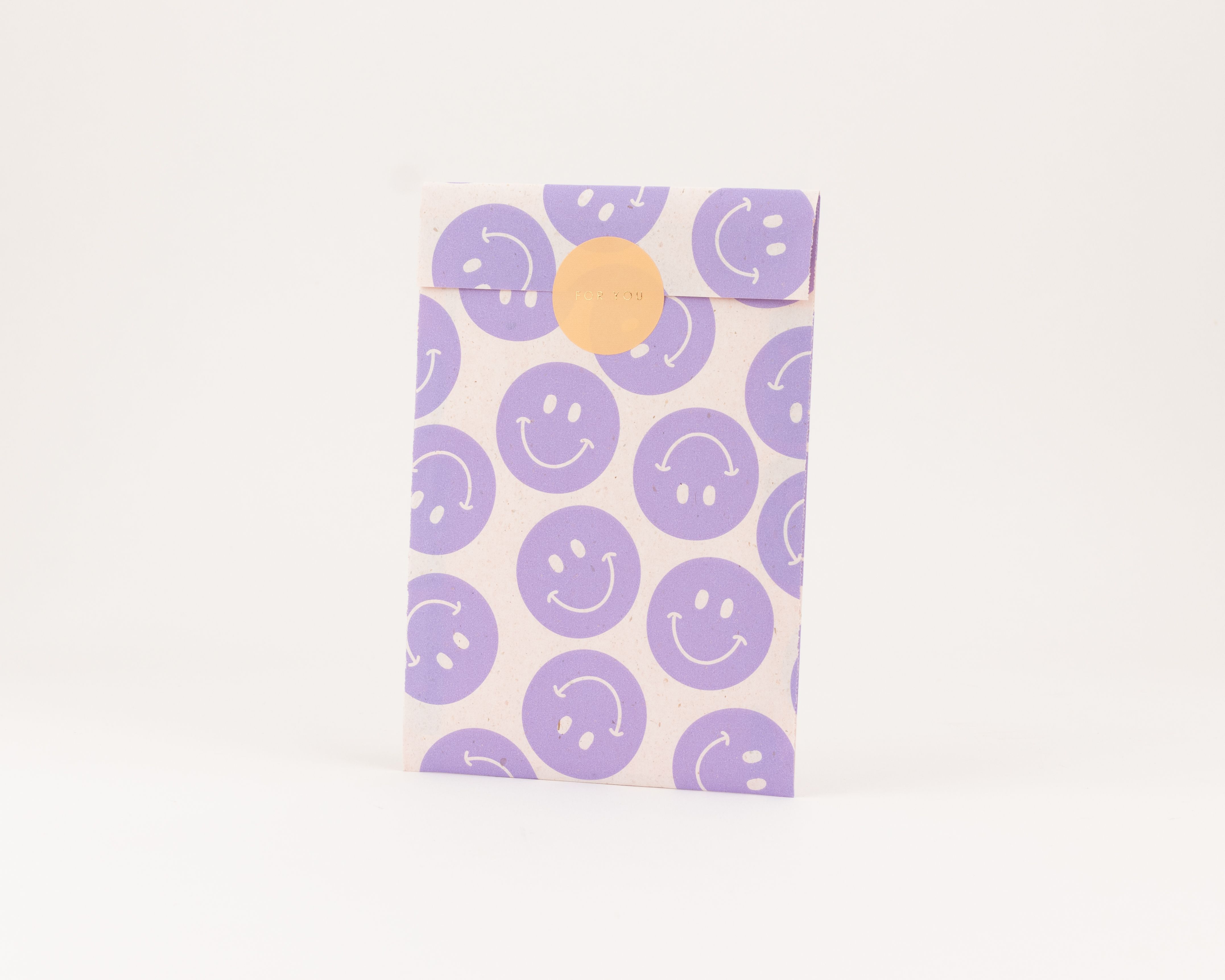 Flachbeutel mit fliederfarbenen Smileys auf hellem Papier und einer passenden Innenseite, verschlossen mit einem orangefarbenen Sticker