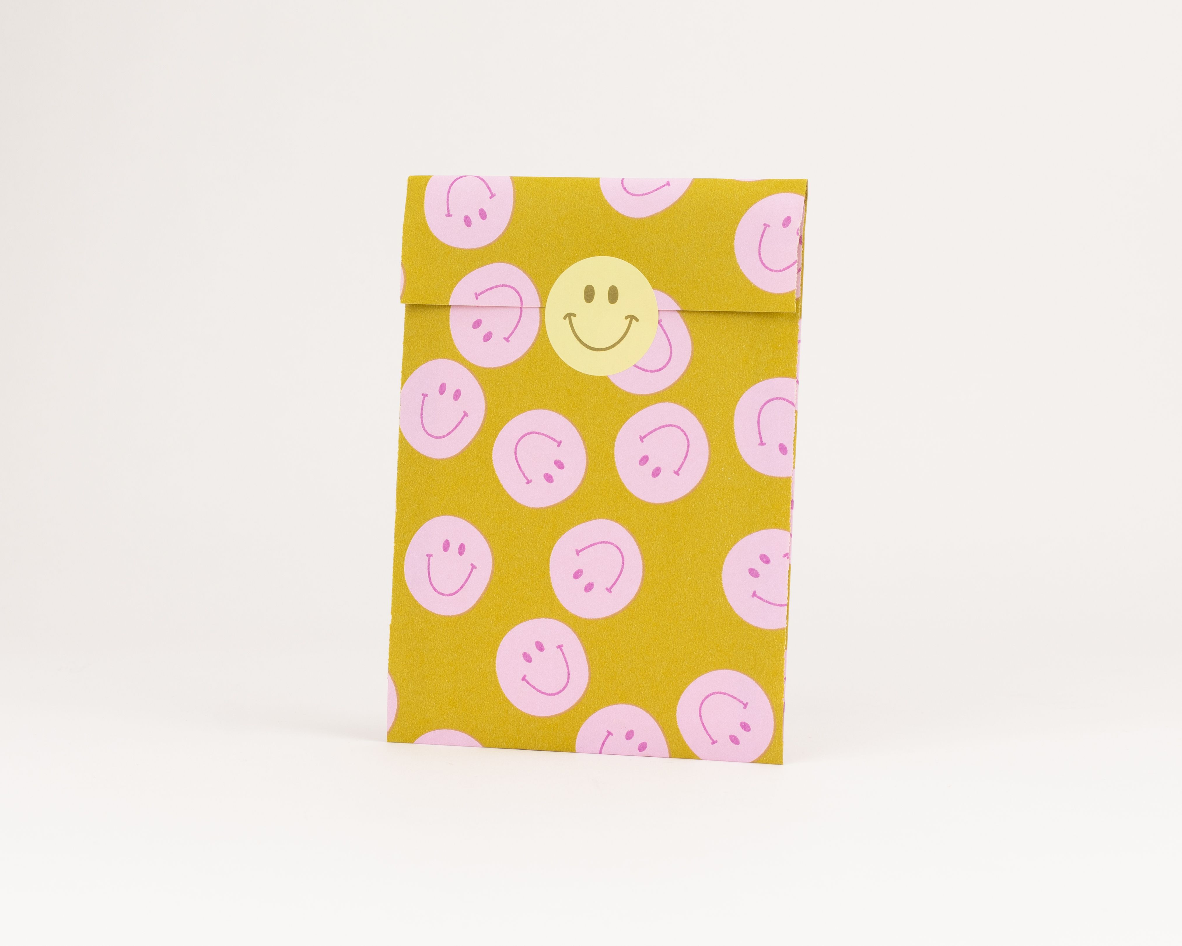 Flachbeutel mit gelber Außenseite, bedruckt mit rosa Smiley-Mustern, verschlossen durch einen gelben runden Smiley-Sticker