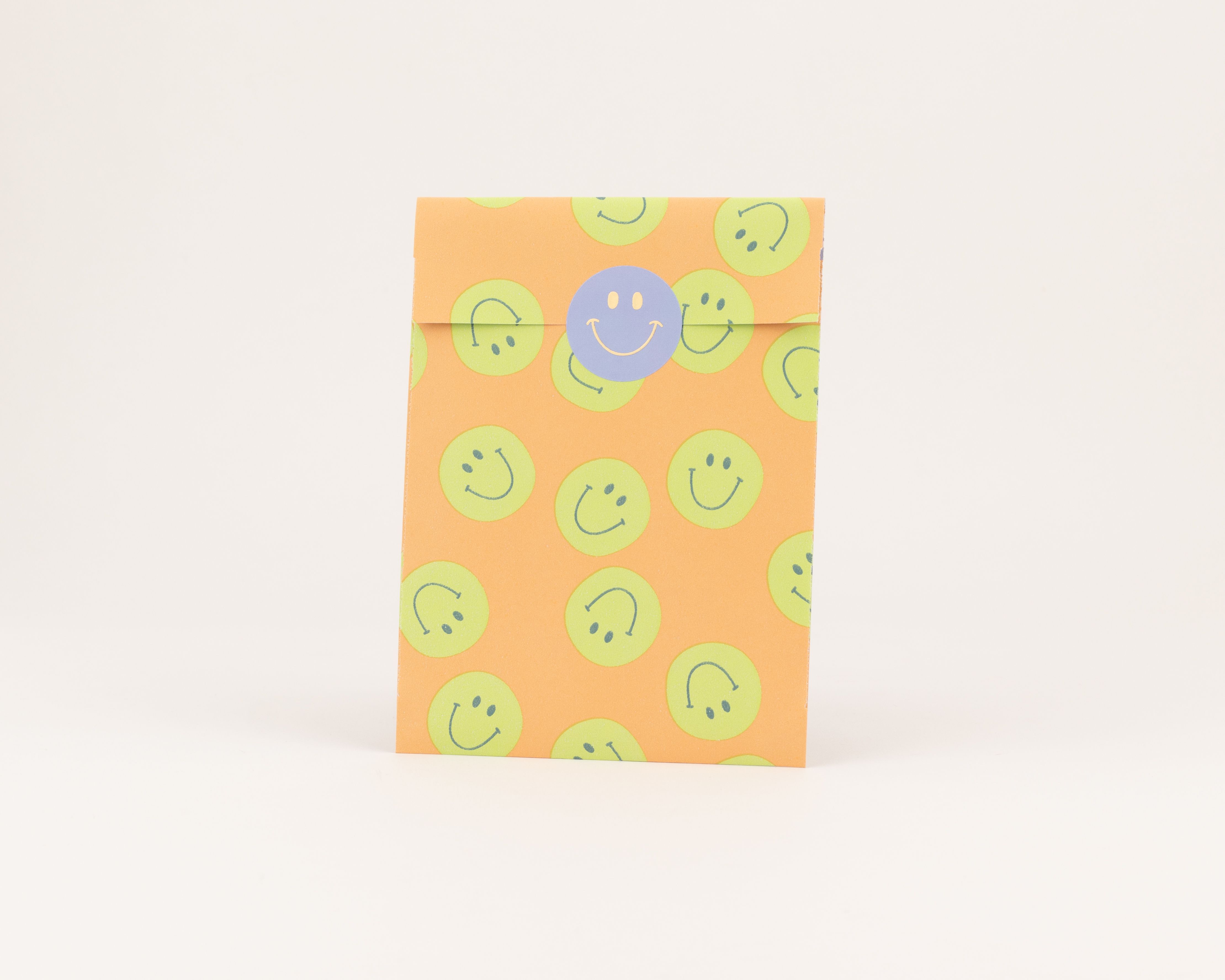 Flachbeutel in Orange mit einem Muster aus gruenen Smileys. Ein runder blauer Aufkleber verschliesst die Tuete. Die Innenseite ist einfarbig Orange.