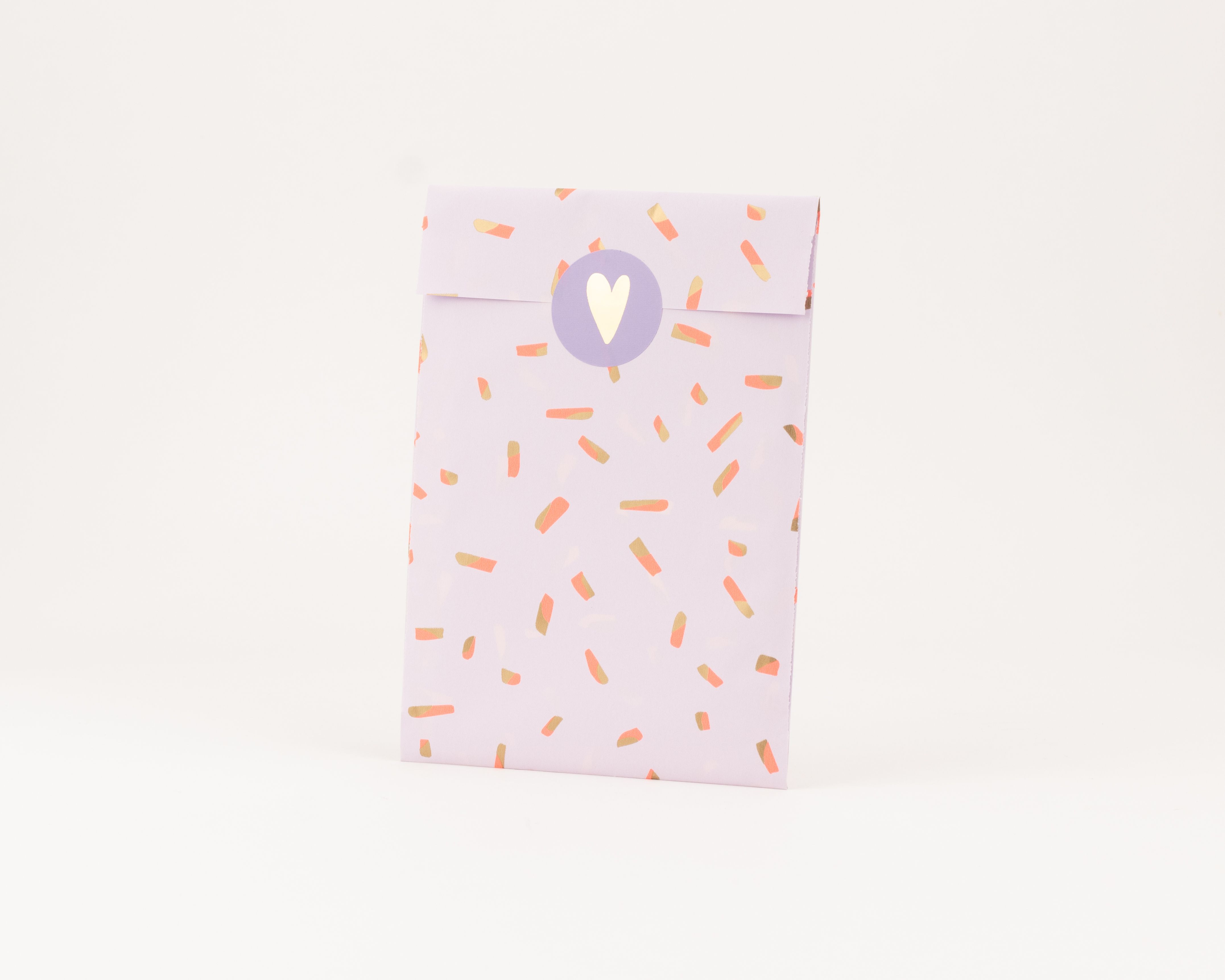 Flachbeutel in hellem Flieder mit Sprinkles-Muster in Koralle und Goldeffekt, verziert mit einem runden Aufkleber und goldenem Herz