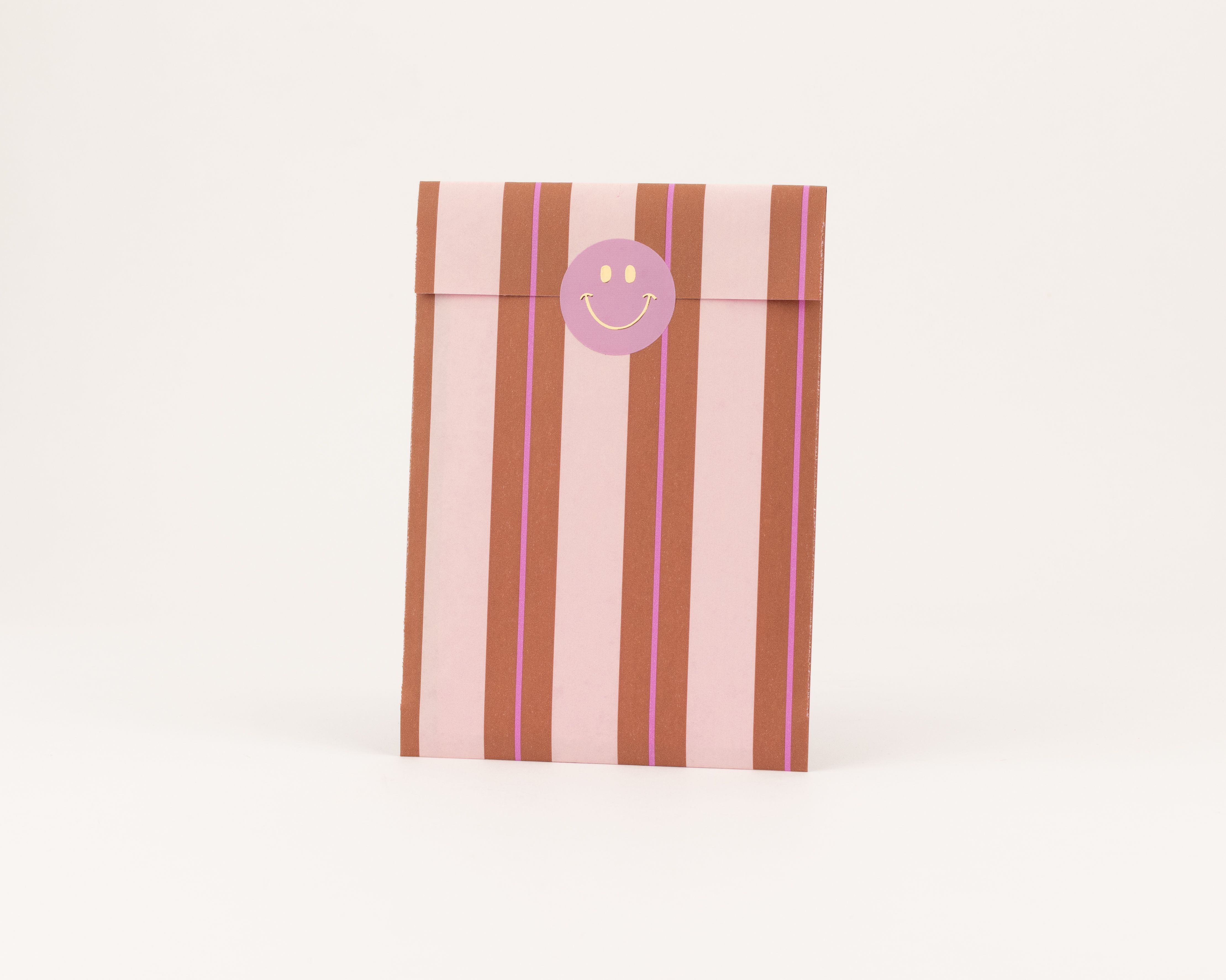 Flachbeutel mit Streifen in Hellrosa und Braun, dazu lila Akzente. Die Innenseite zeigt ebenfalls Streifen und einen lila Smiley-Sticker