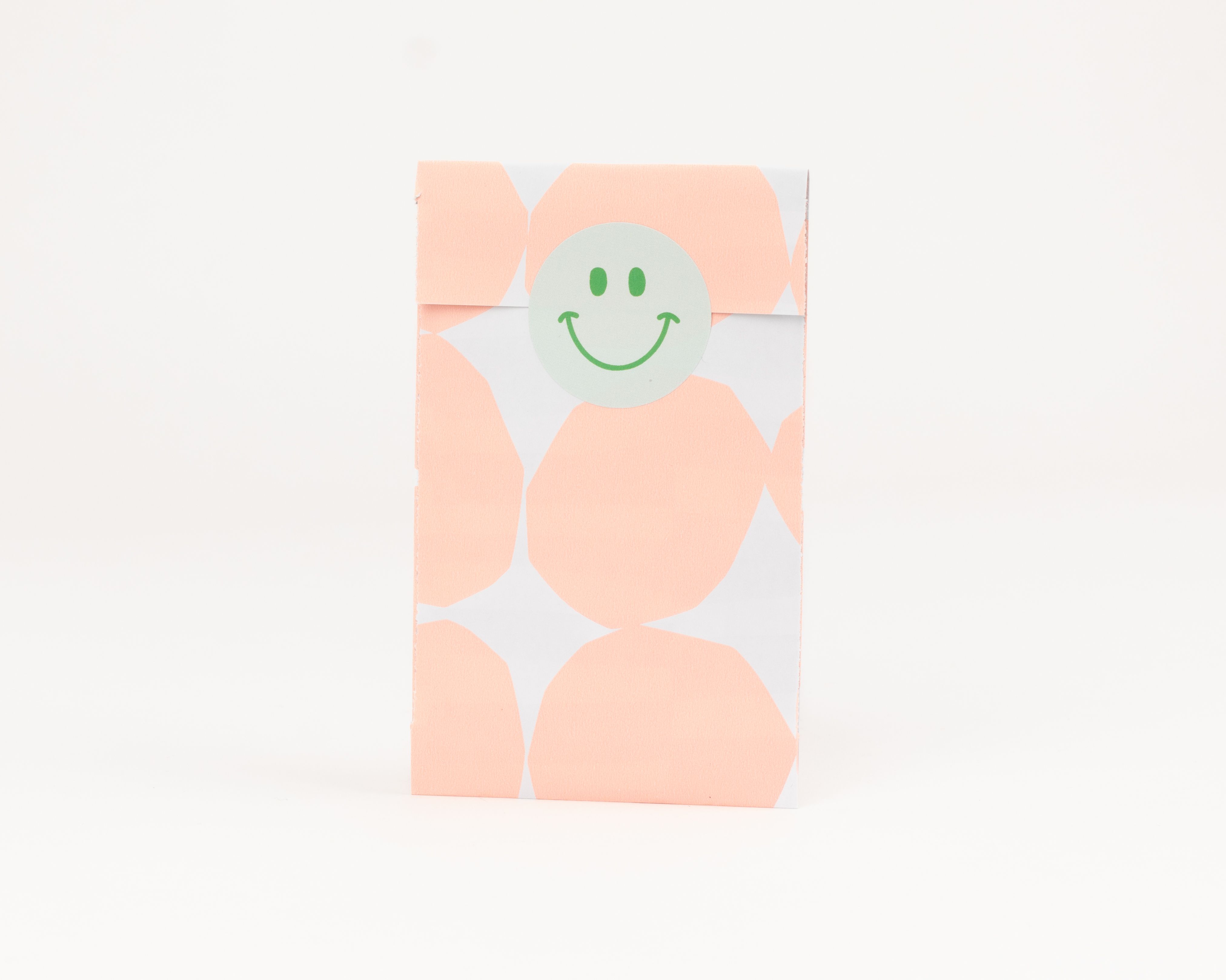 Flachbeutel mit pfirsichfarbenen Punkten auf weißem Grund und weißer Innenseite. Ein hellgrüner Smiley Aufkleber verschließt den Beutel