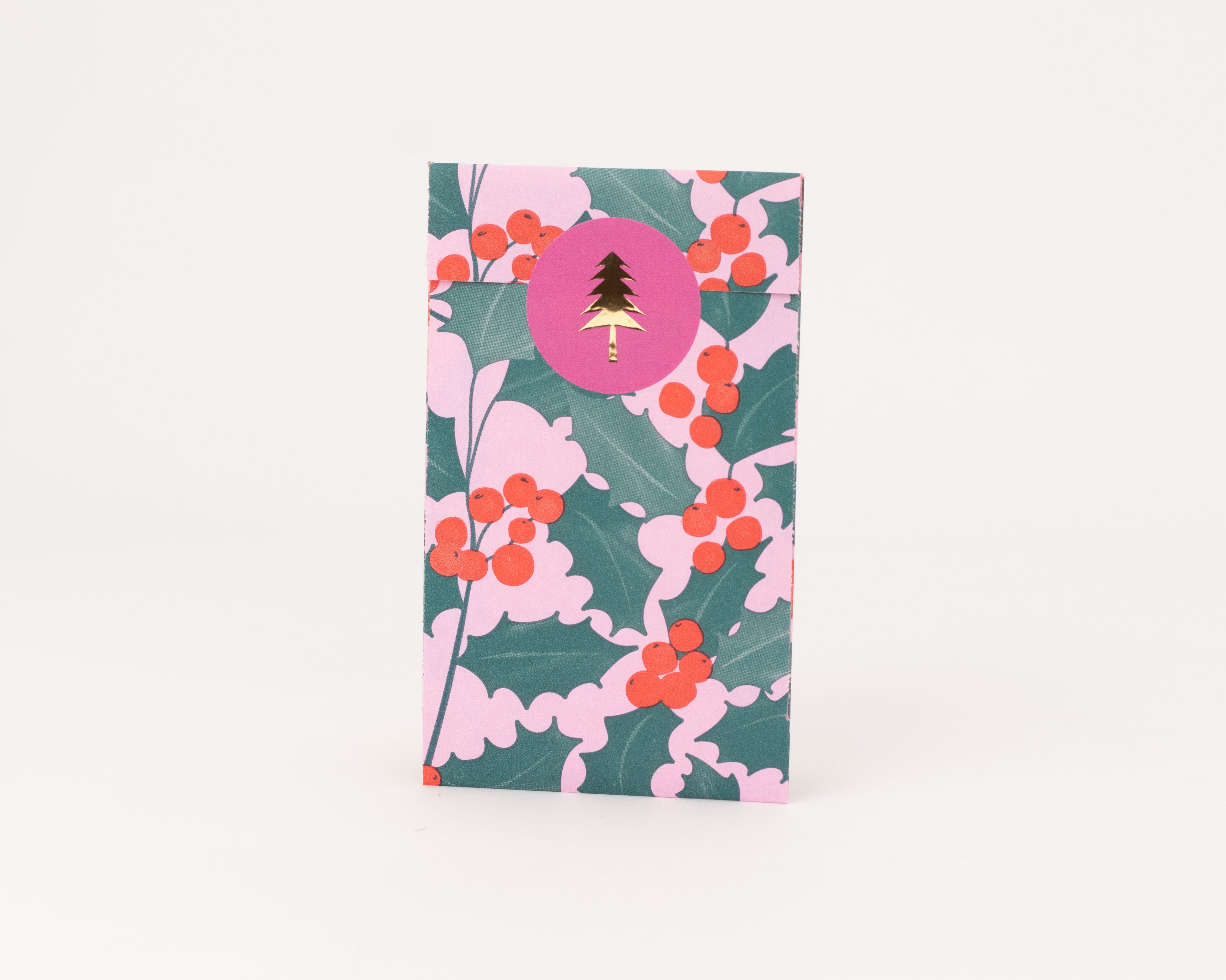 Flachbeutel mit Ilex-Muster: gruene Blaetter, rote Beeren auf rosa Grund. Rosa Aufkleber mit goldenem Tannenbaum. Innenseite rosa