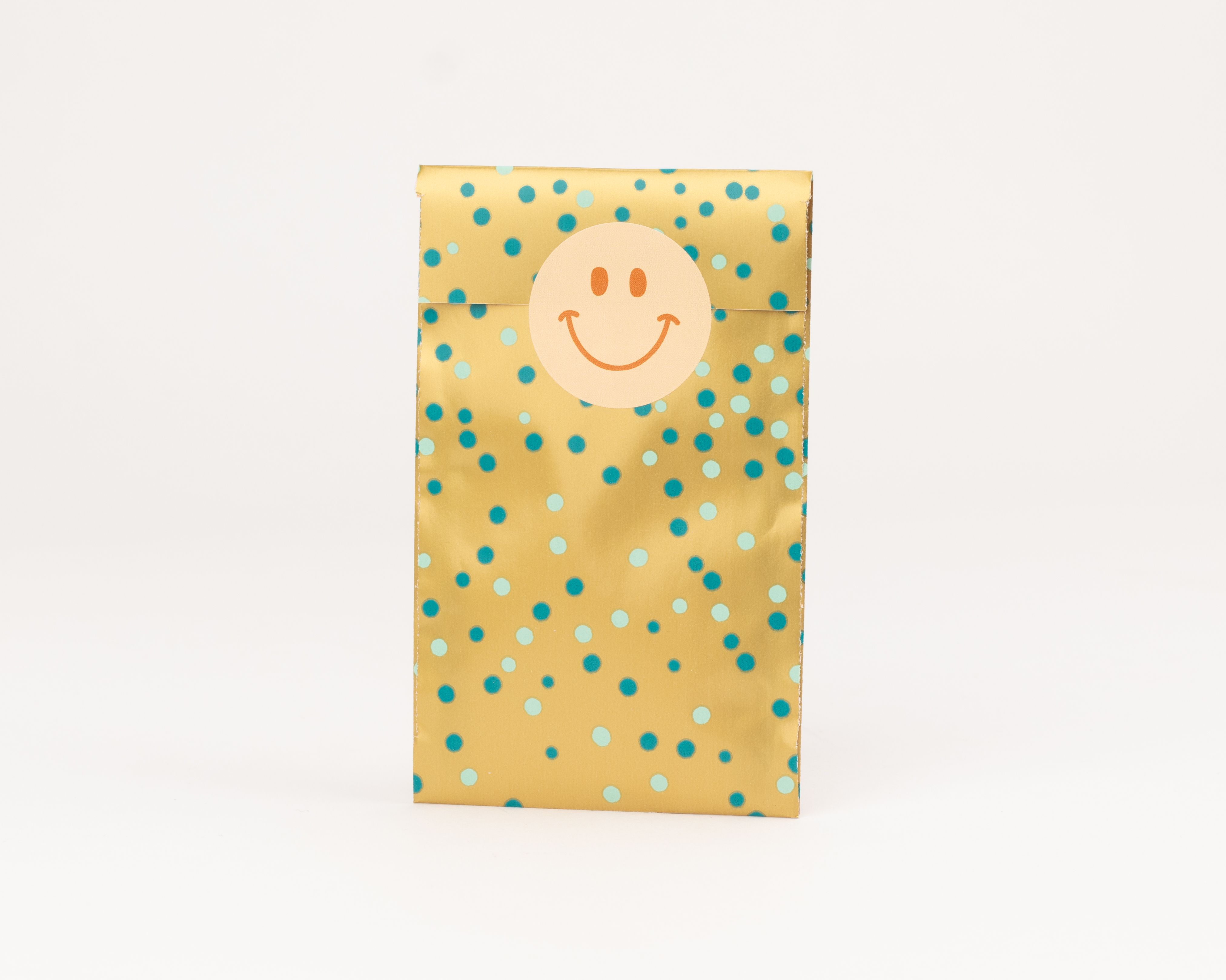 Flachbeutel in Gold mit blauen und hellblauen Konfettipunkten Beigefarbener Smiley-Sticker und dunkelbraune Innenseite sind sichtbar