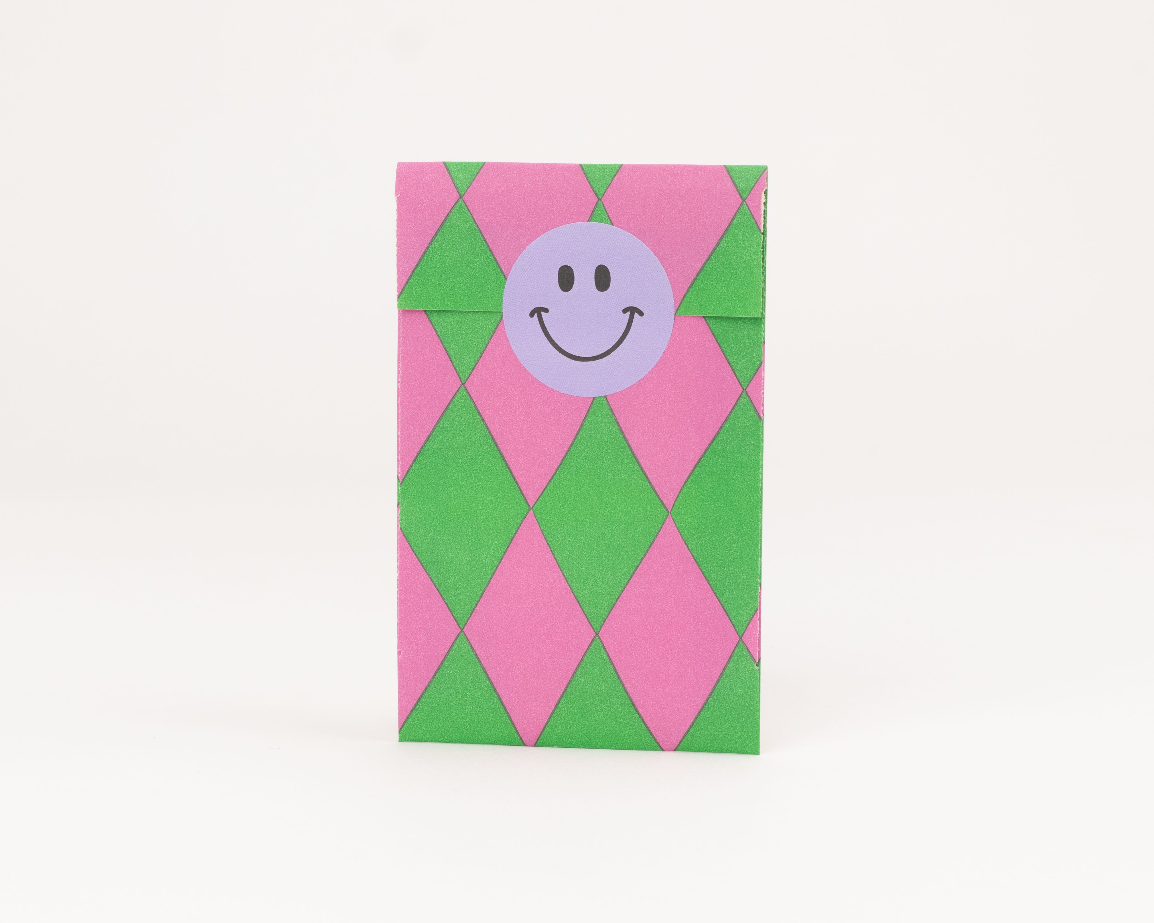 Flachbeutel mit pinken und grünen Rauten, verziert mit einem runden lila Smiley Aufkleber zum Verschließen