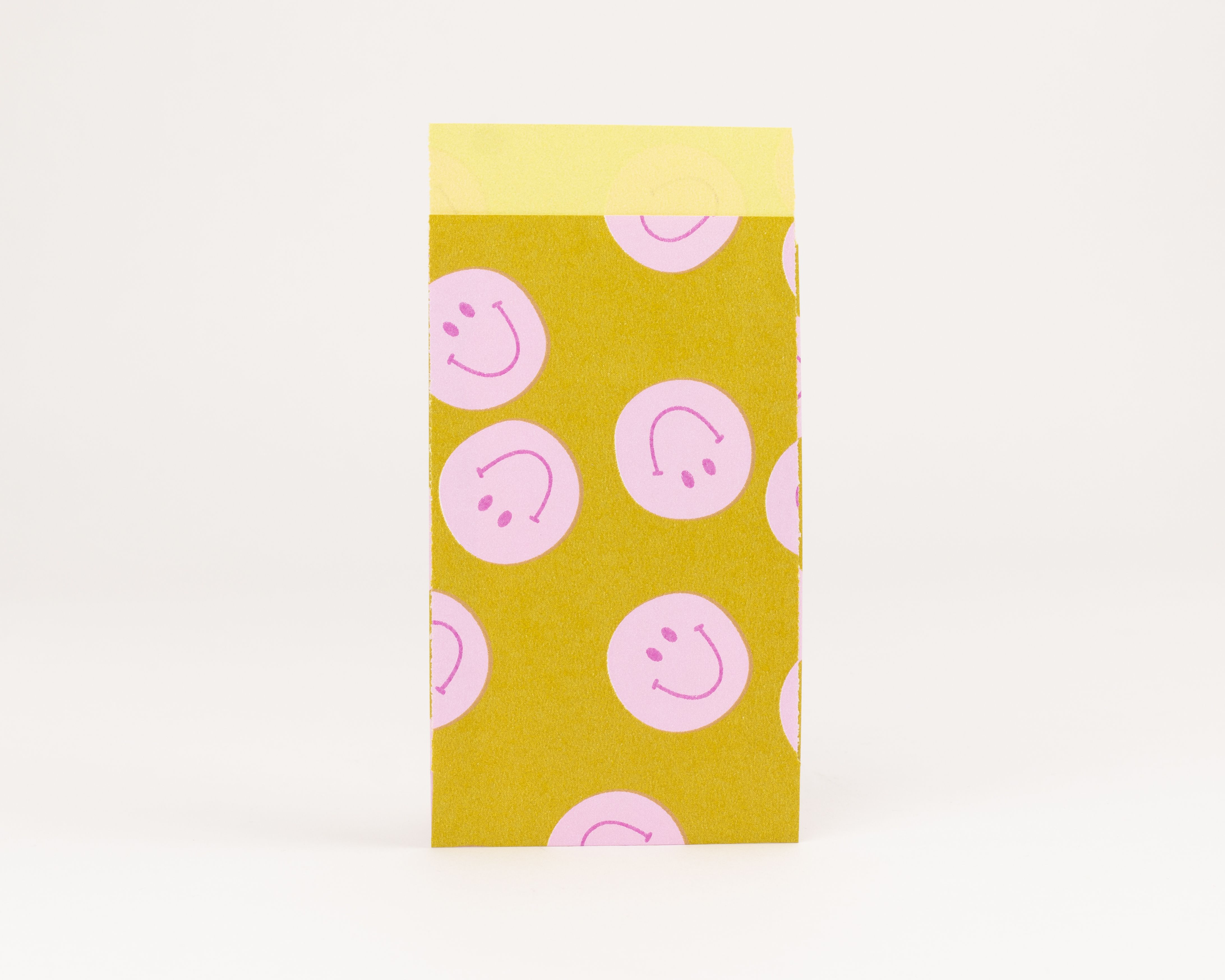 Flachbeutel mit gelb-olivefarbenem Hintergrund und rosa lachenden Smiley-Gesichtern, die Innenseite ist hellgelb