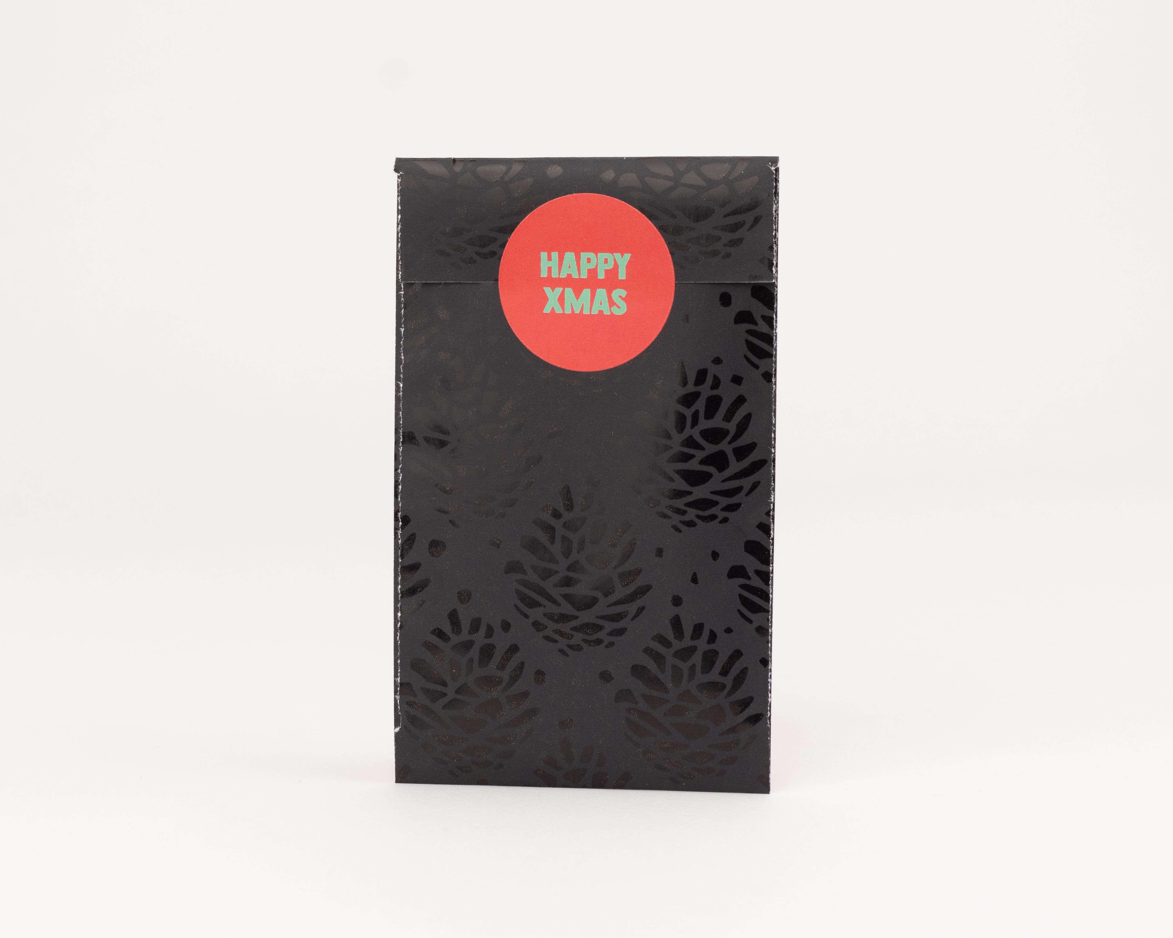 Flachbeutel sind schwarz mit einem glaenzenden Tannenzapfen-Muster, verschlossen mit einem runden roten Aufkleber mit gruenem Happy Xmas Schriftzug