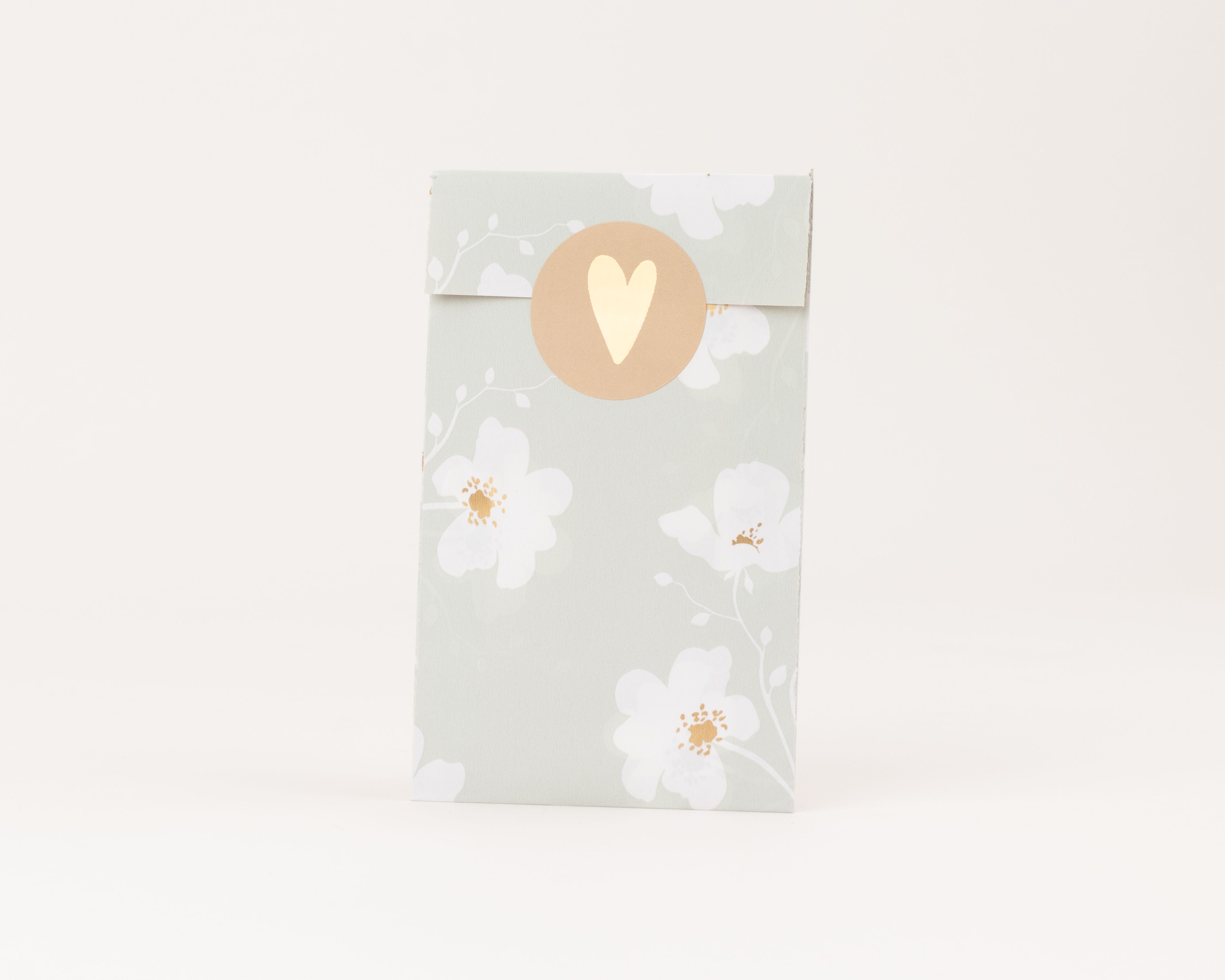 Flachbeutel in Salbeigrün mit weißen Wiesenblüten und Goldeffekten. Ein runder Aufkleber mit einem goldenen Herz auf ockerfarbenem Grund