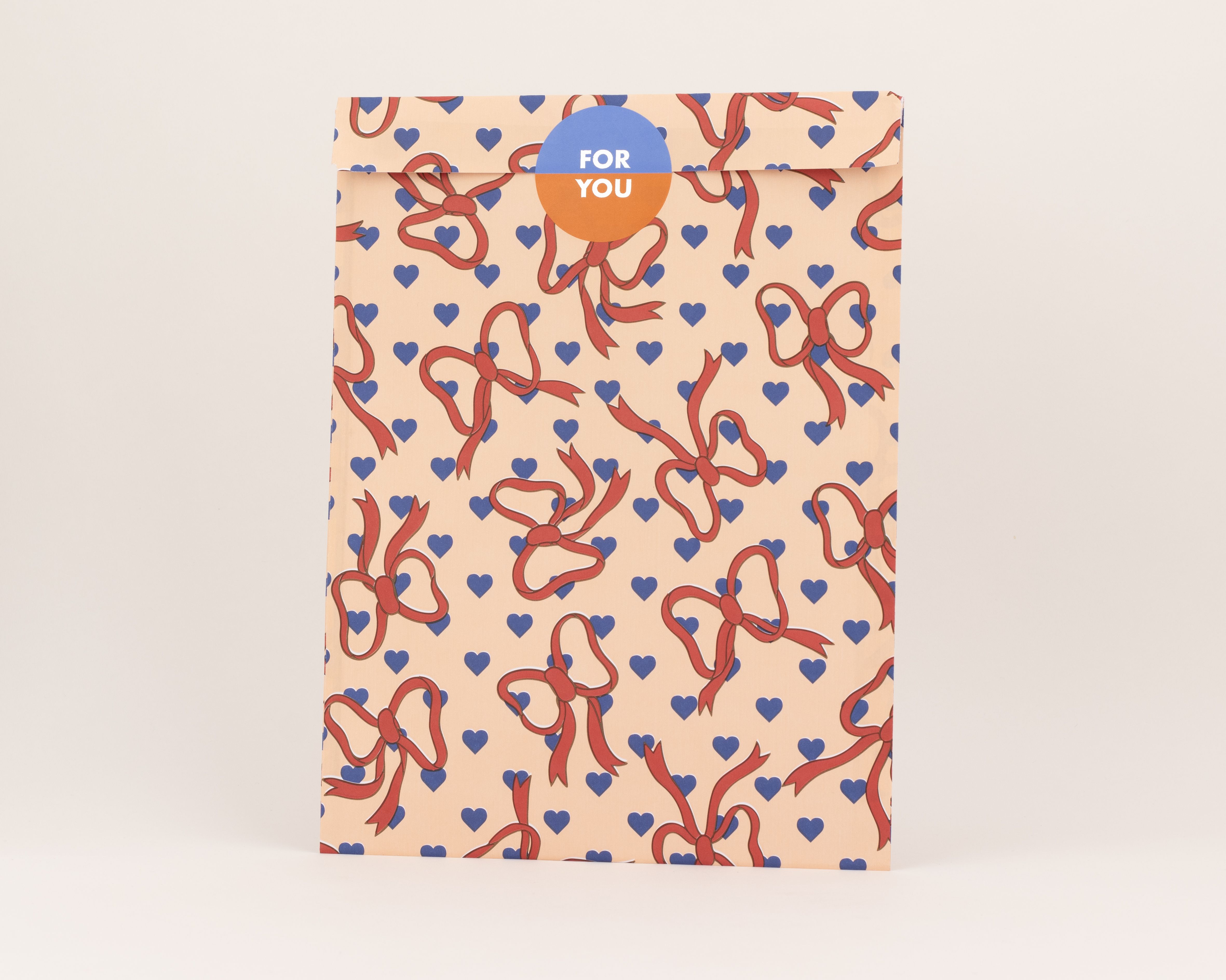 Flachbeutel mit einem Muster aus roten Schleifen und kornblumenblauen Herzen auf pfirsichfarbenem Hintergrund