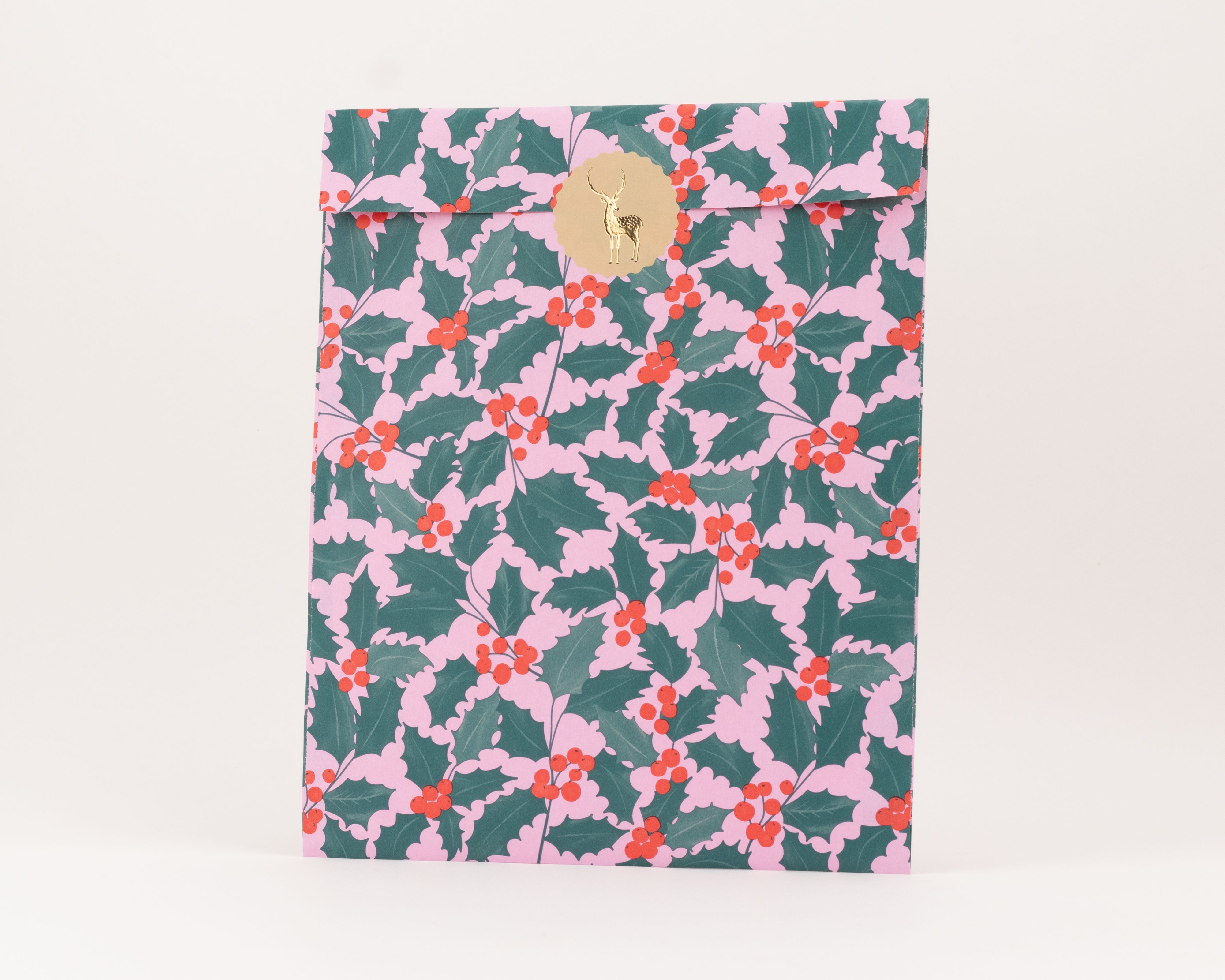 Flachbeutel mit rosa Hintergrund und grünem Ilex-Muster, verziert mit roten Beeren und einem goldenen Aufkleber mit Rentier-Motiv