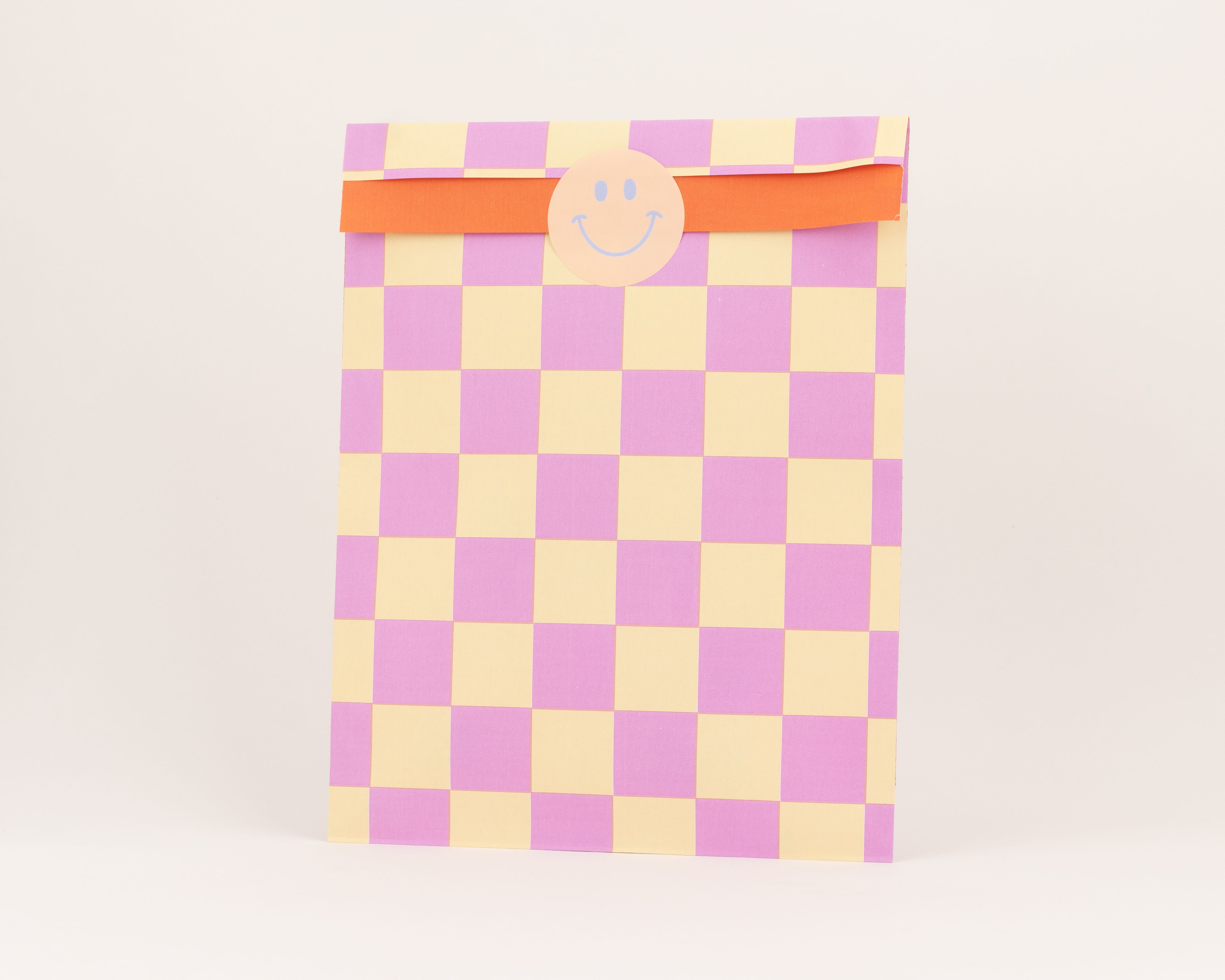 Flachbeutel mit Schachbrettmuster in Pink und Gelb, die Innenseite des Umschlags ist orange, ein runder Smiley-Aufkleber ziert die Vorderseite