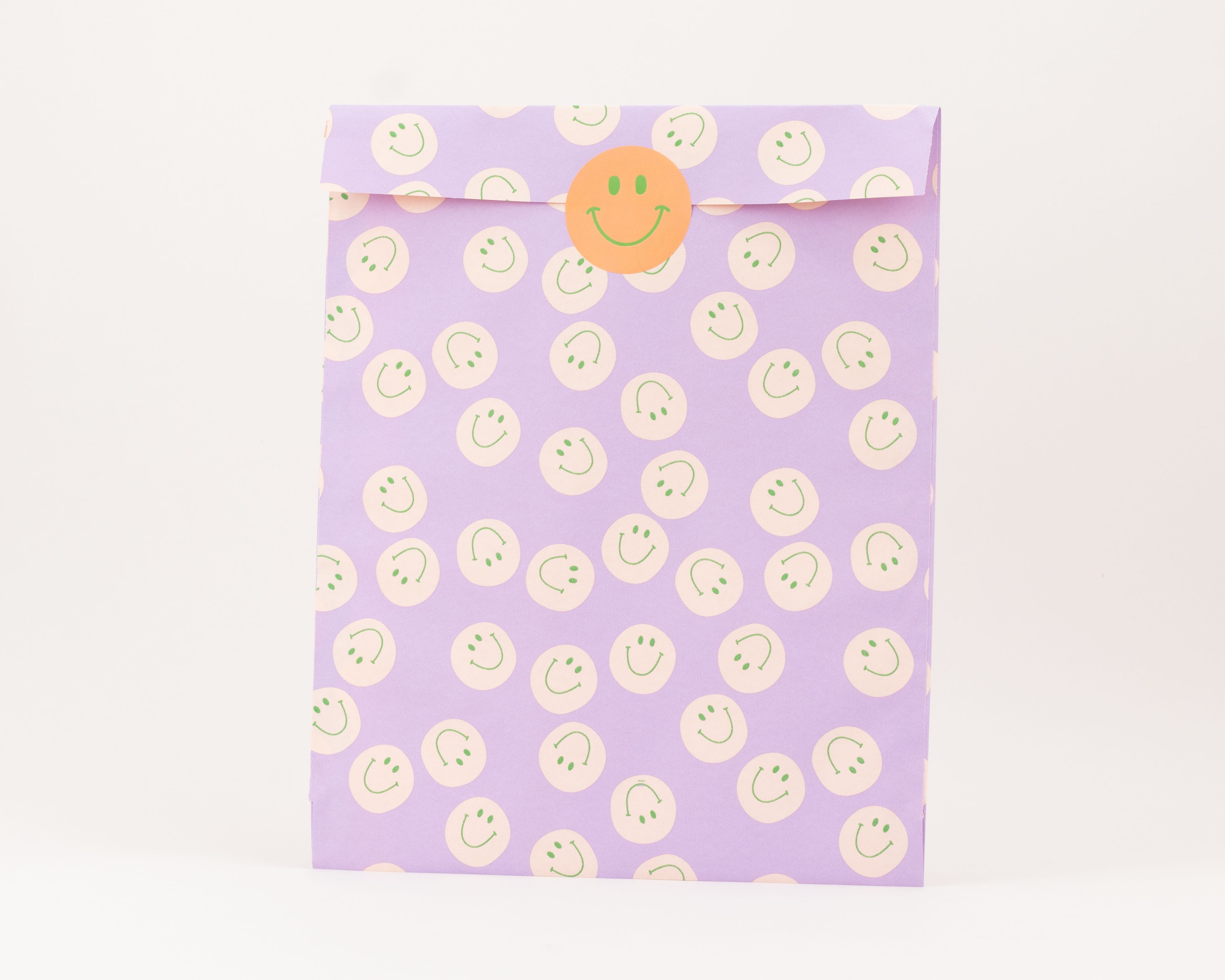 Flachbeutel in Flieder mit elfenbeinfarbenen Kreisen und gruenen Smileys, verschlossen mit einem runden orangefarbenen Smiley-Aufkleber