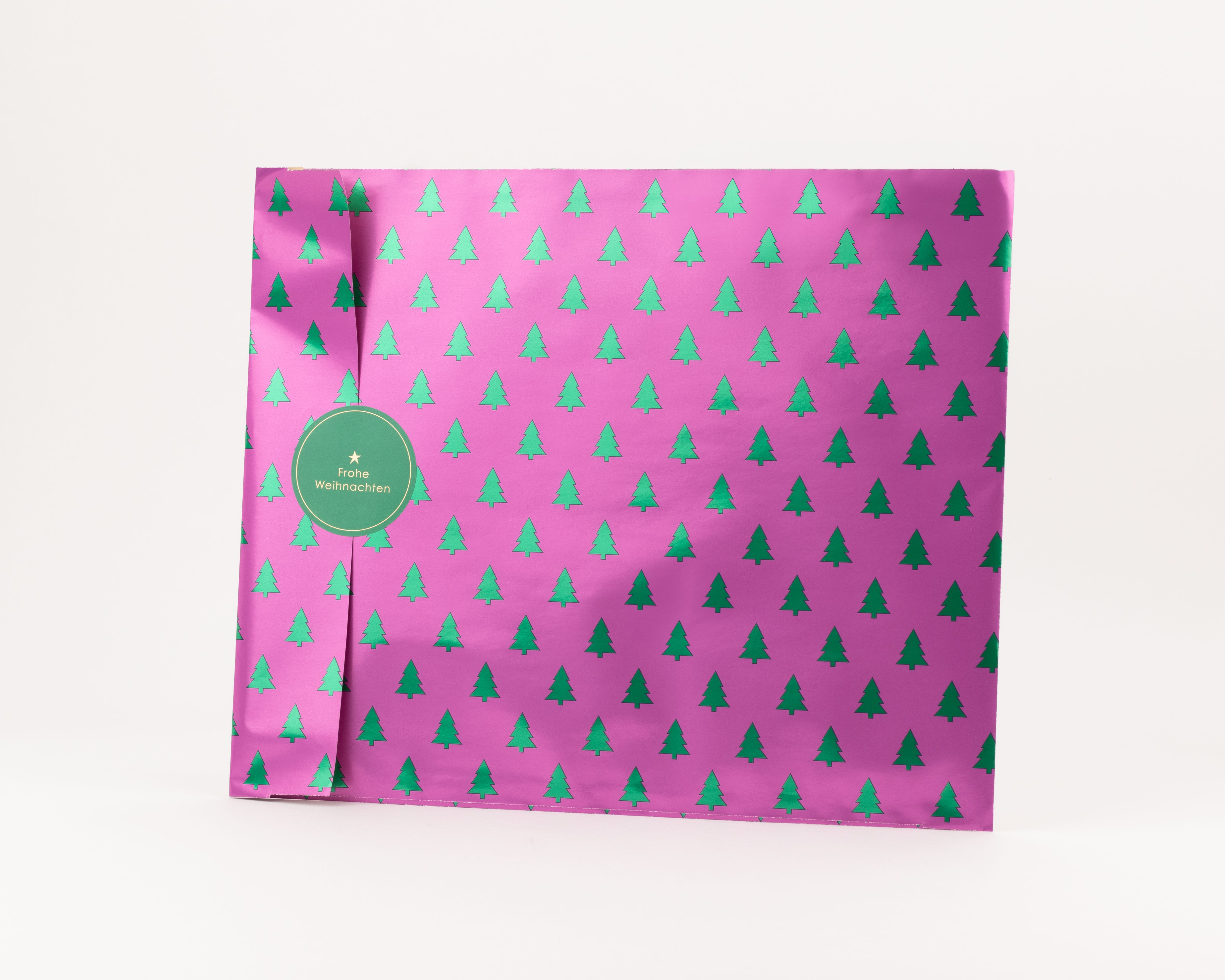 Flachbeutel mit einem pinken Hintergrund und einem wiederkehrenden Muster aus metallisch gruenen Tannenbaeumen, wobei die Innenseite beige ist