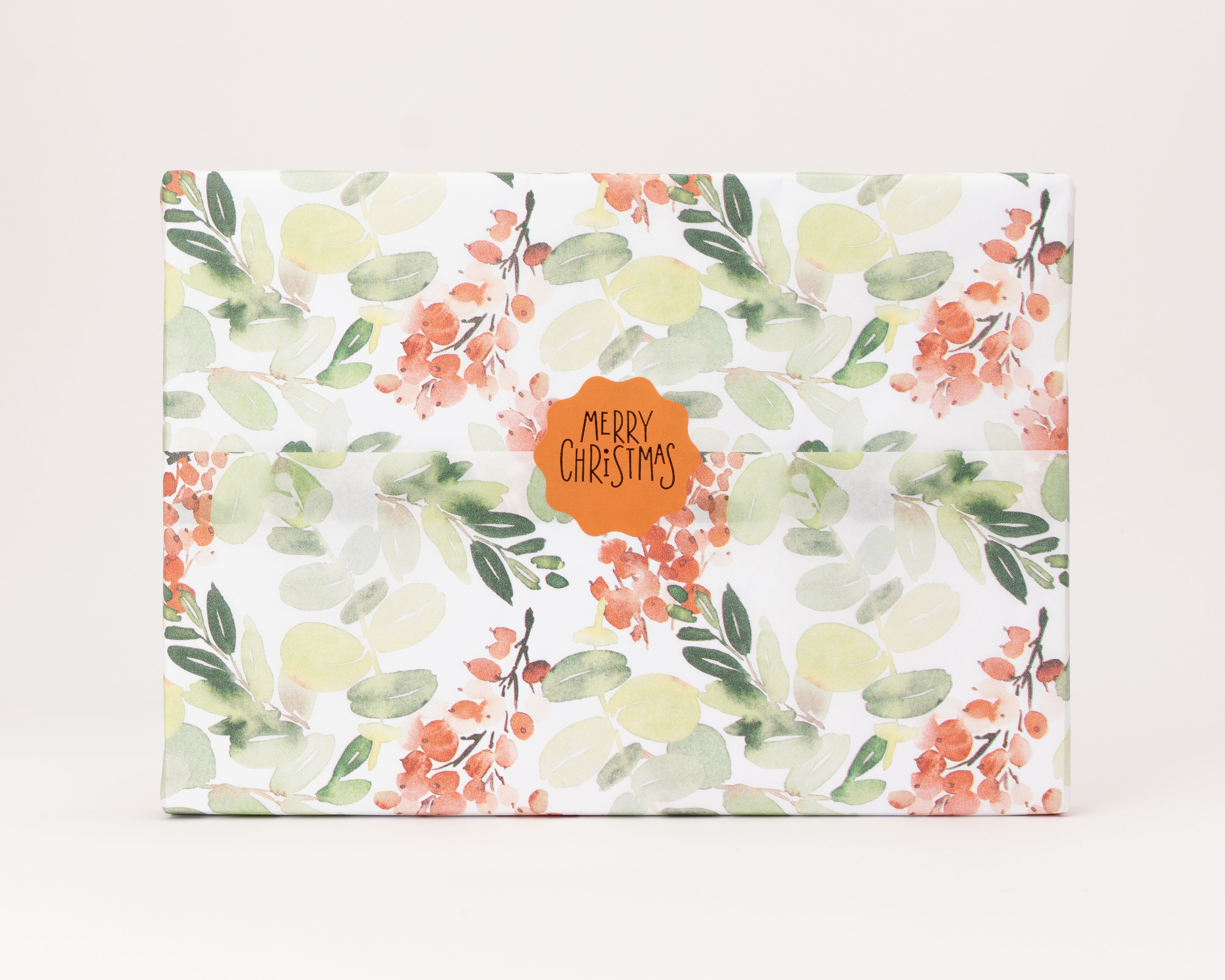 Seidenpapier mit Beerenzweigen-Muster in Grün und Orange auf weißem Hintergrund. Darauf ein orangefarbener Aufkleber 'MERRY CHRISTMAS'