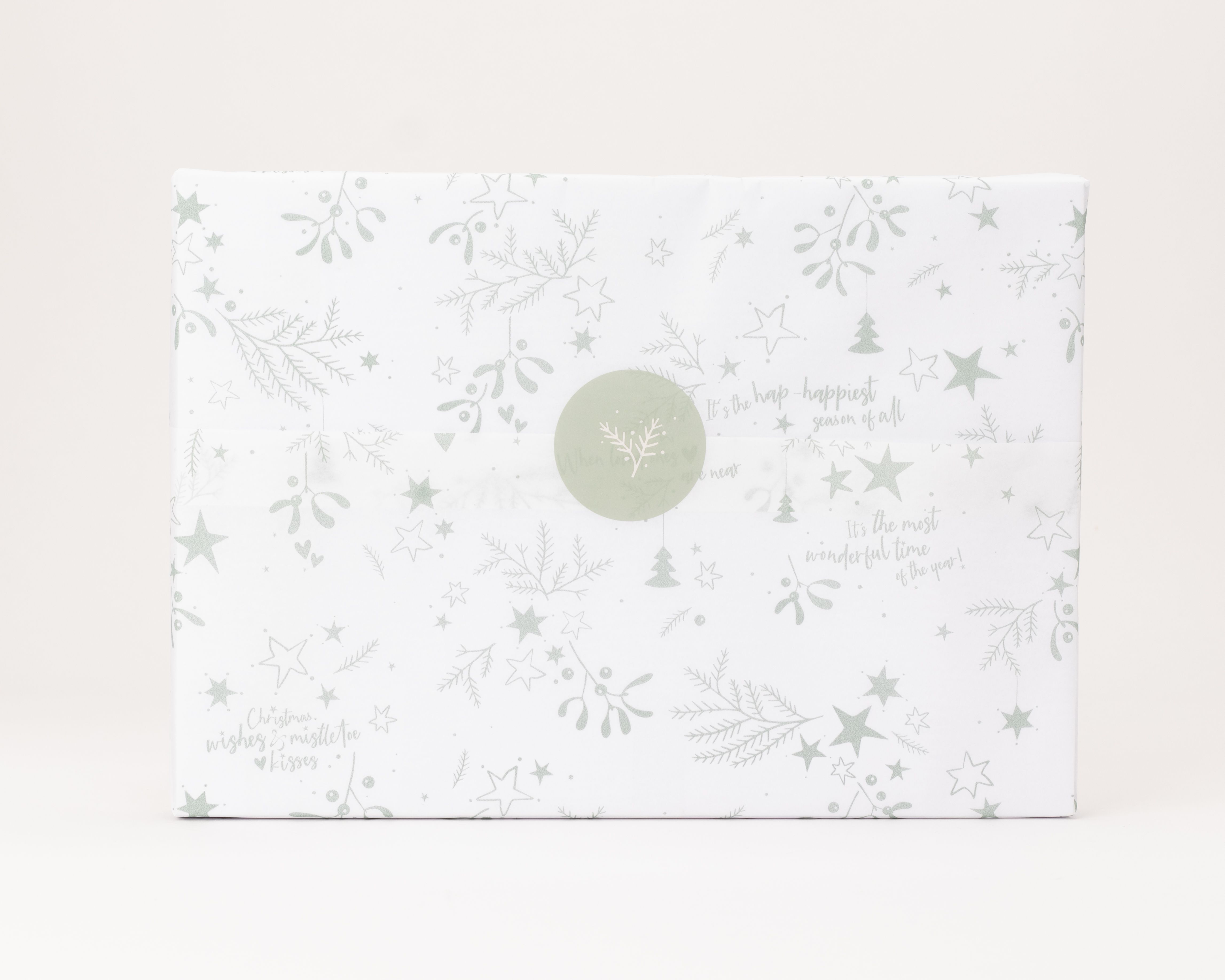 Seidenpapier ist weiß, mit salbeigrünen Weihnachtsmotiven und englischen Wunschtexten gemustert. Darauf ein runder, salbeigrüner Aufkleber mit weißem Zweigmuster