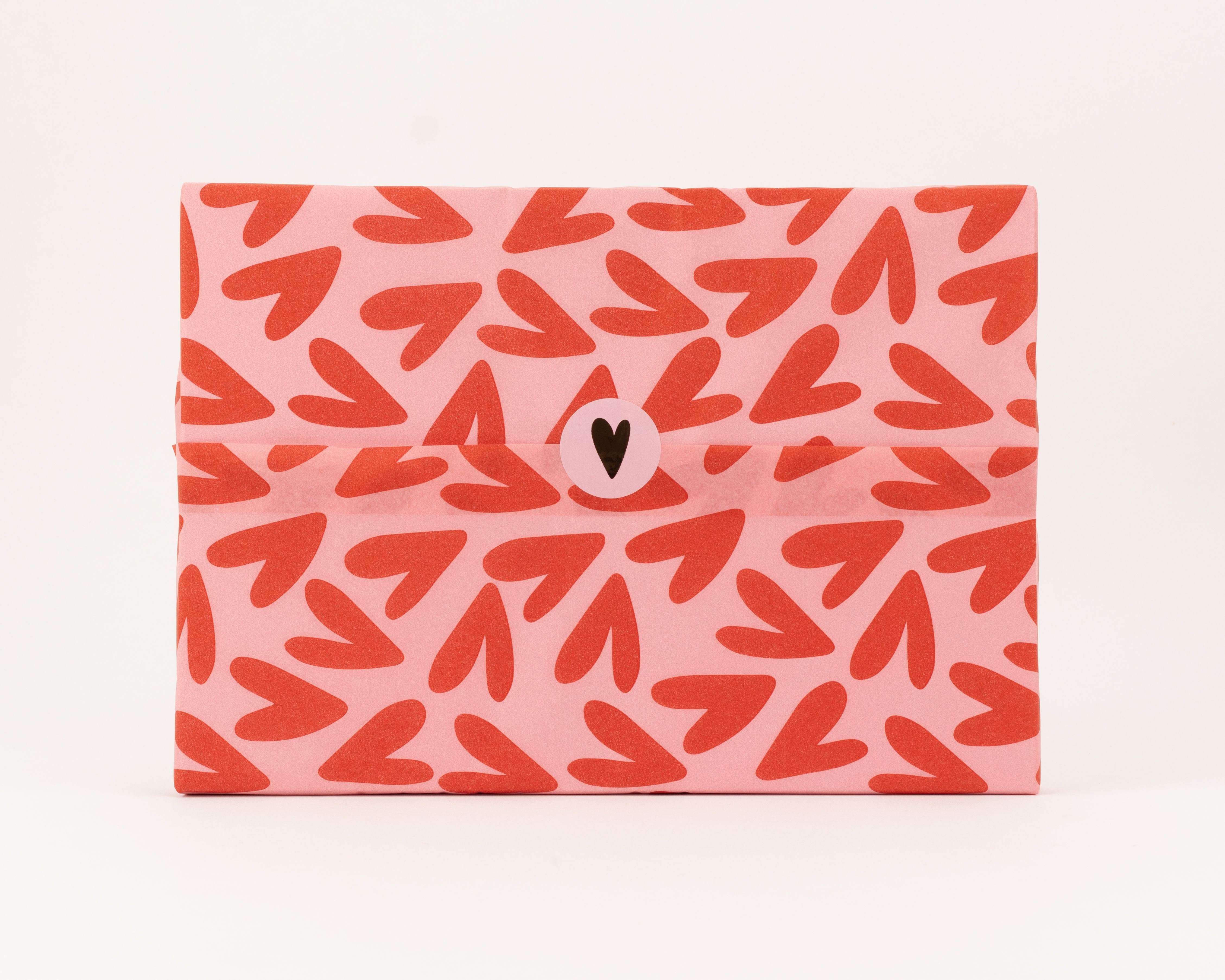 Seidenpapier mit einem Muster aus roten Herzen auf rosa Hintergrund. Darauf ein schwarzer herzförmiger Aufkleber